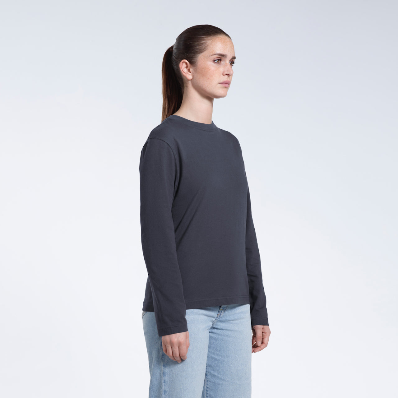A woman looking away from the camera with her arms by her sides, modelling a plain dark grey long sleeve organic cotton t shirt.