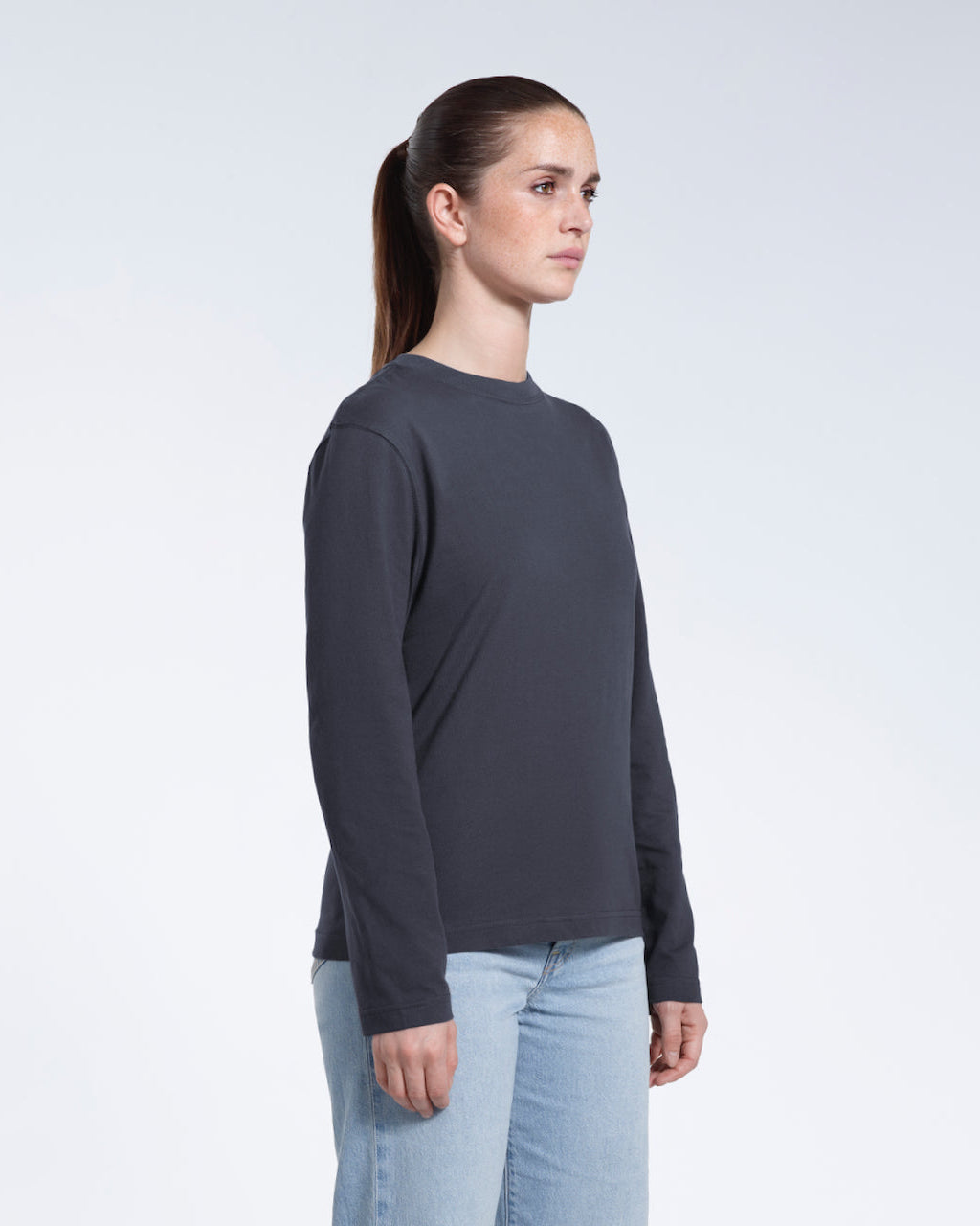 A woman looking away from the camera with her arms by her sides, modelling a plain dark grey long sleeve organic cotton t shirt.