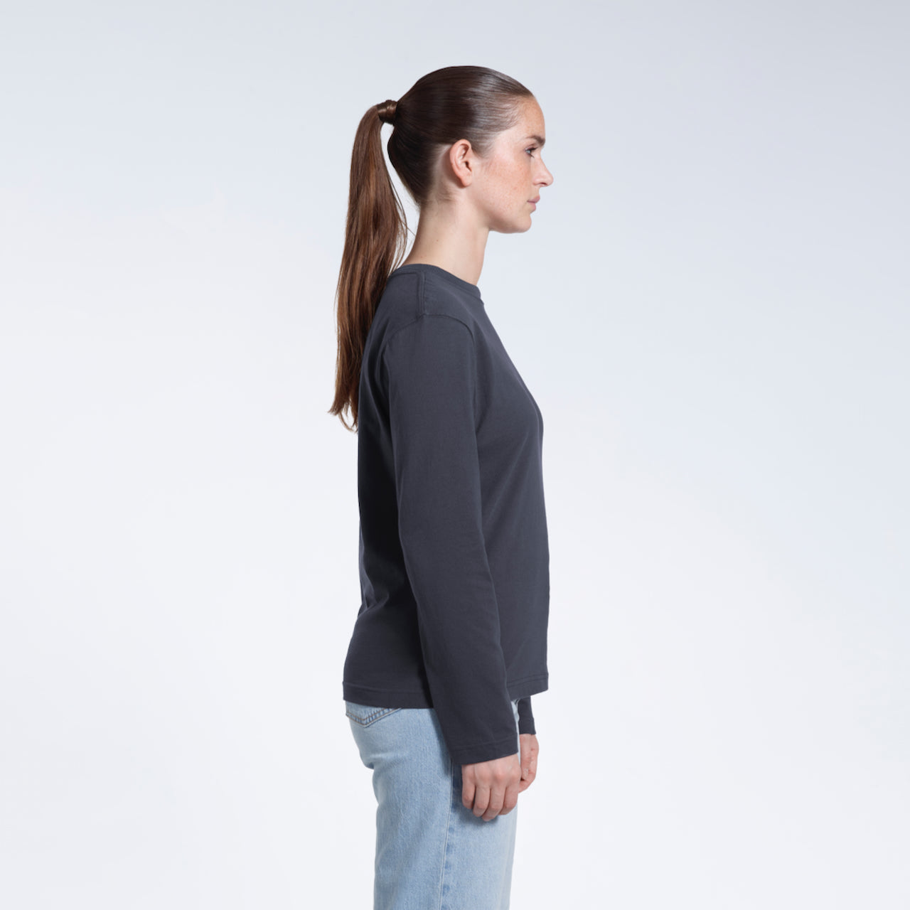 A woman facing side on with her arms by her sides, modelling a plain dark grey long sleeve organic cotton t shirt.