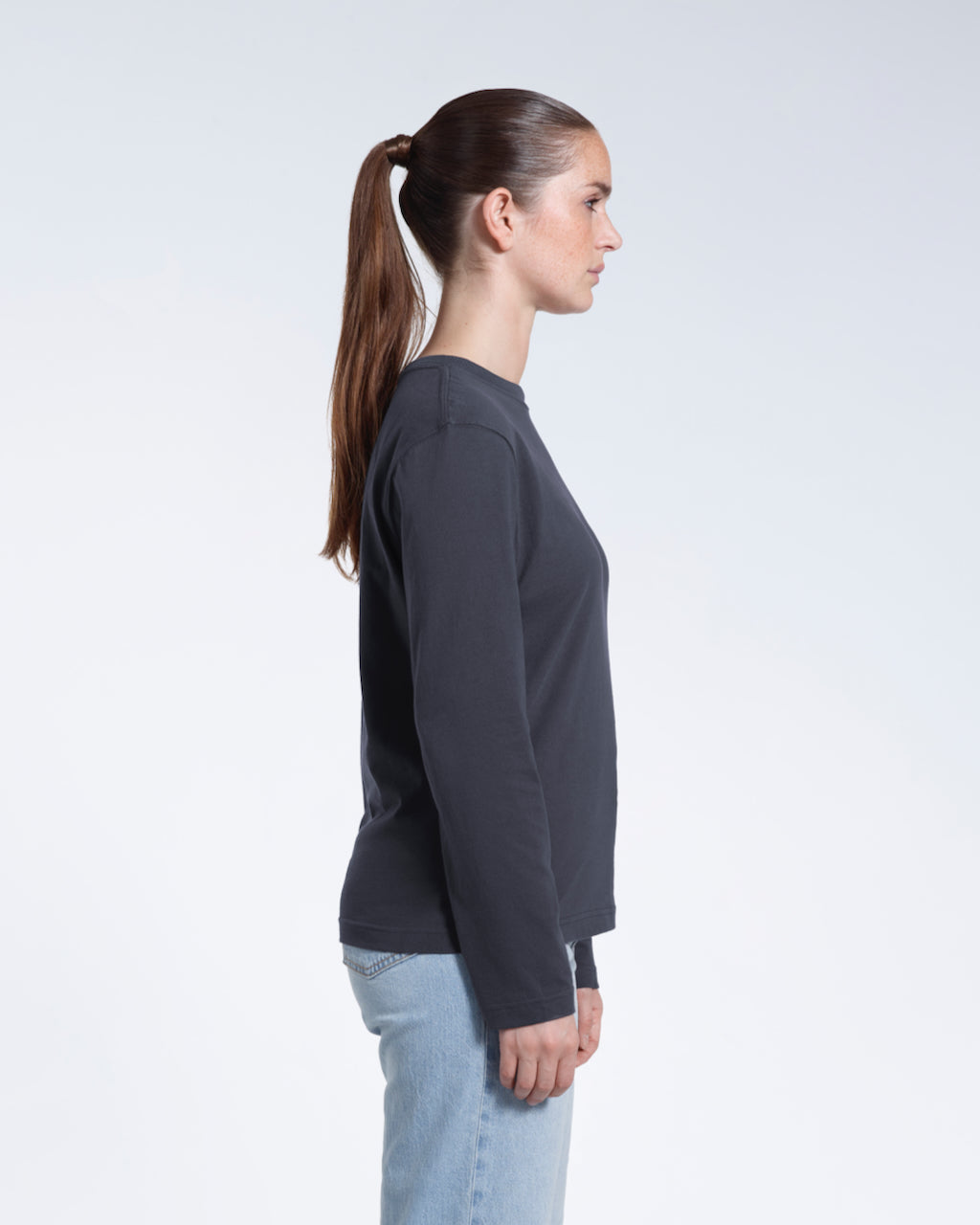 A woman facing side on with her arms by her sides, modelling a plain dark grey long sleeve organic cotton t shirt.