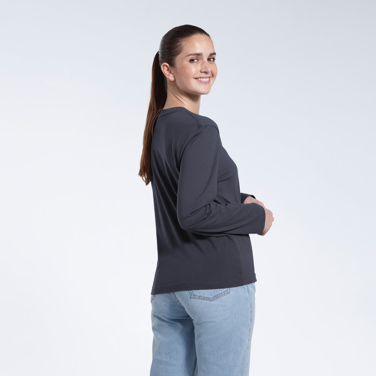 A woman turned round smiling into the camera, modelling a plain dark grey long sleeve organic cotton t shirt.