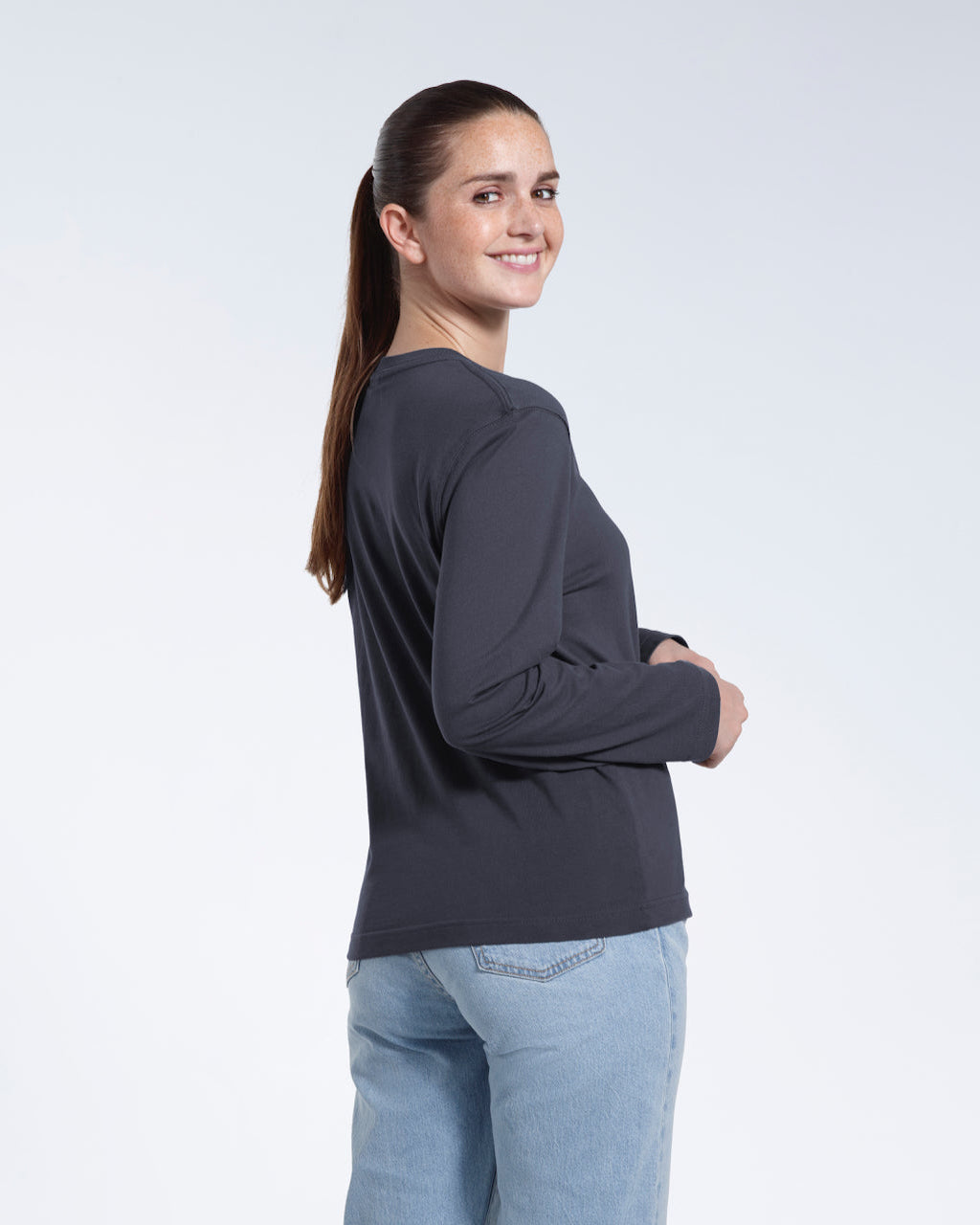 A woman turned round smiling into the camera, modelling a plain dark grey long sleeve organic cotton t shirt.