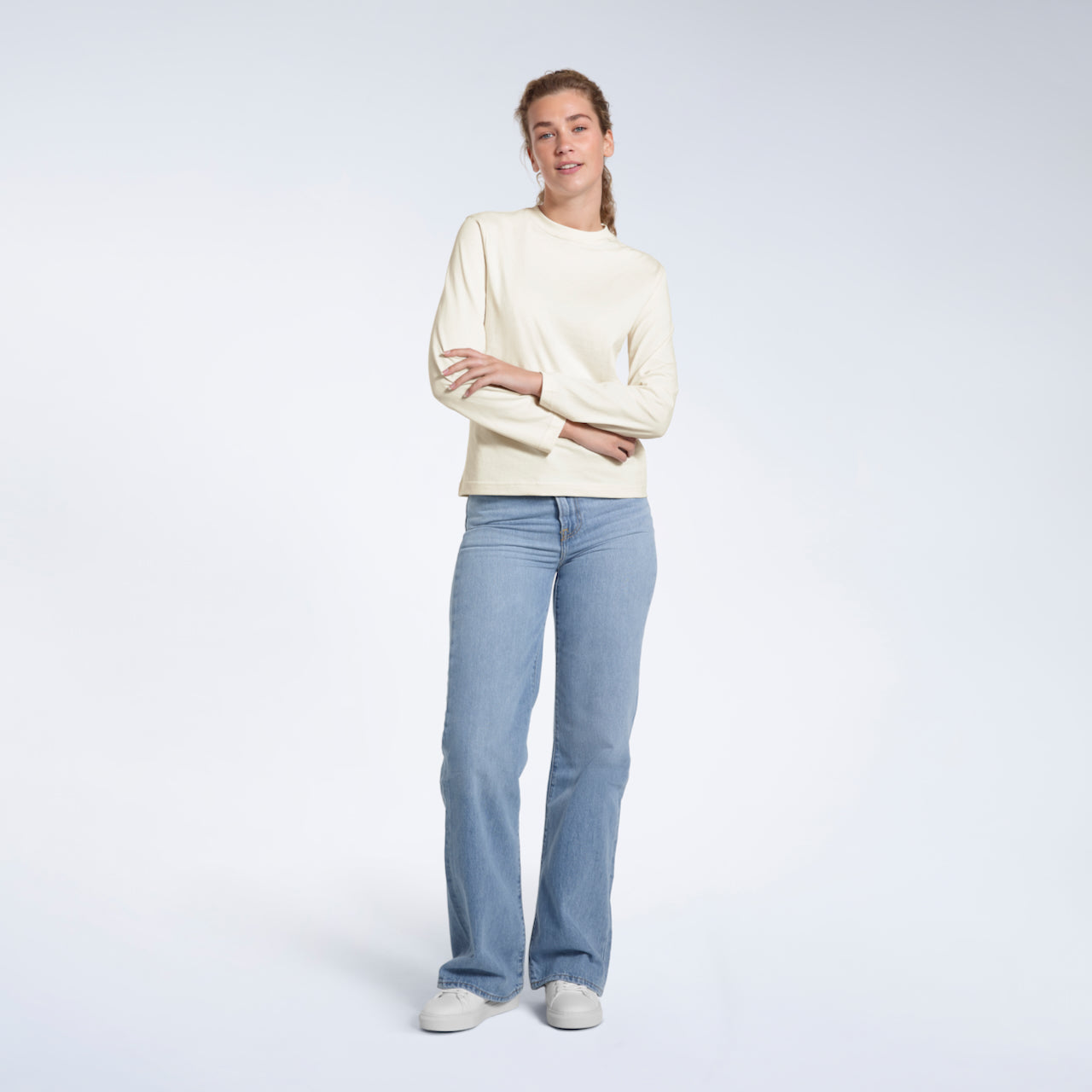 A full body photo of a woman looking into the camera with her arms folded, modelling a plain ecru long sleeve organic cotton t shirt.
