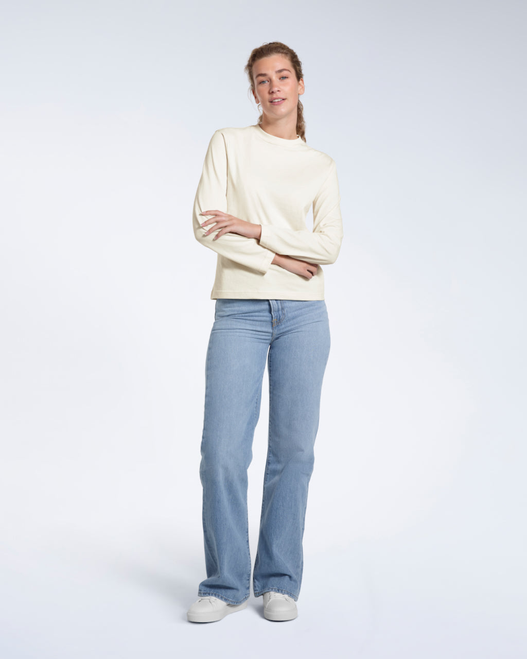 A full body photo of a woman looking into the camera with her arms folded, modelling a plain ecru long sleeve organic cotton t shirt.