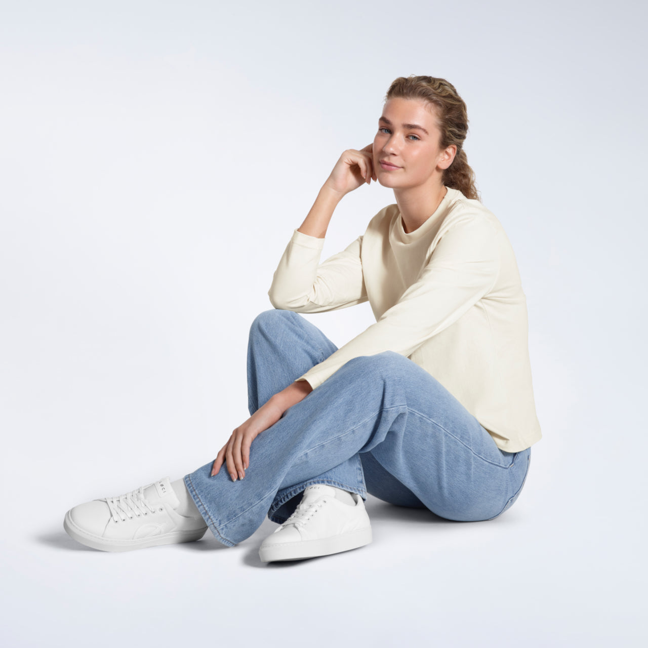 A woman sitting on the floor smiling at the camera, modelling a plain ecru long sleeve organic cotton t shirt.
