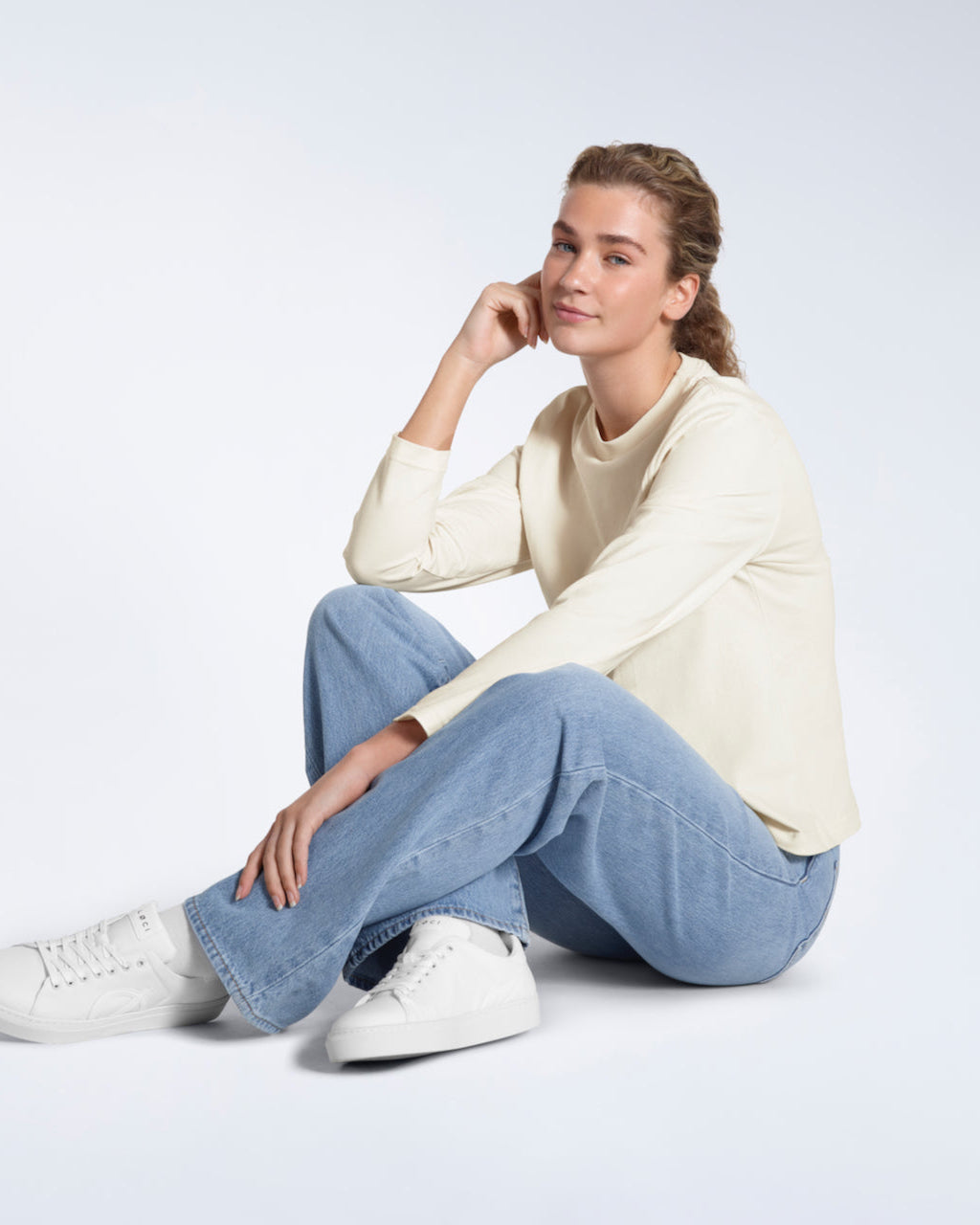 A woman sitting on the floor smiling at the camera, modelling a plain ecru long sleeve organic cotton t shirt.