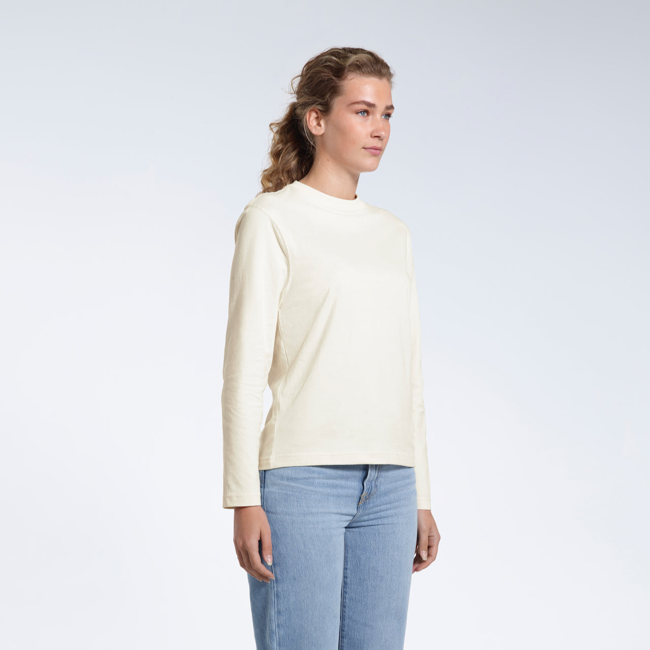 A woman looking away from the camera with her arms by her sides, modelling a plain ecru long sleeve organic cotton t shirt.