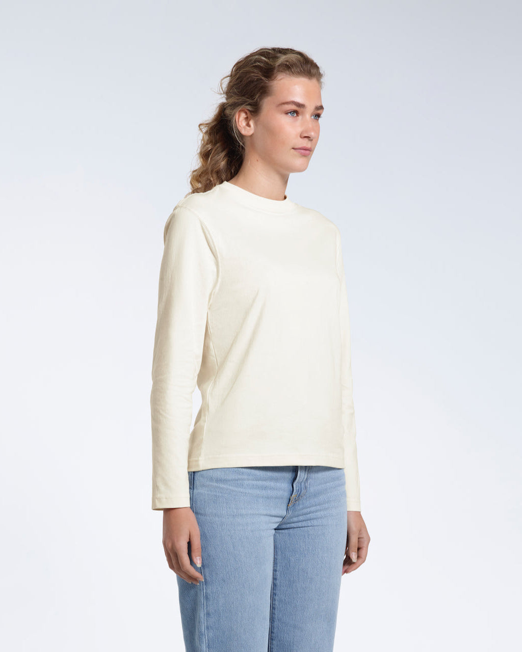 A woman looking away from the camera with her arms by her sides, modelling a plain ecru long sleeve organic cotton t shirt.