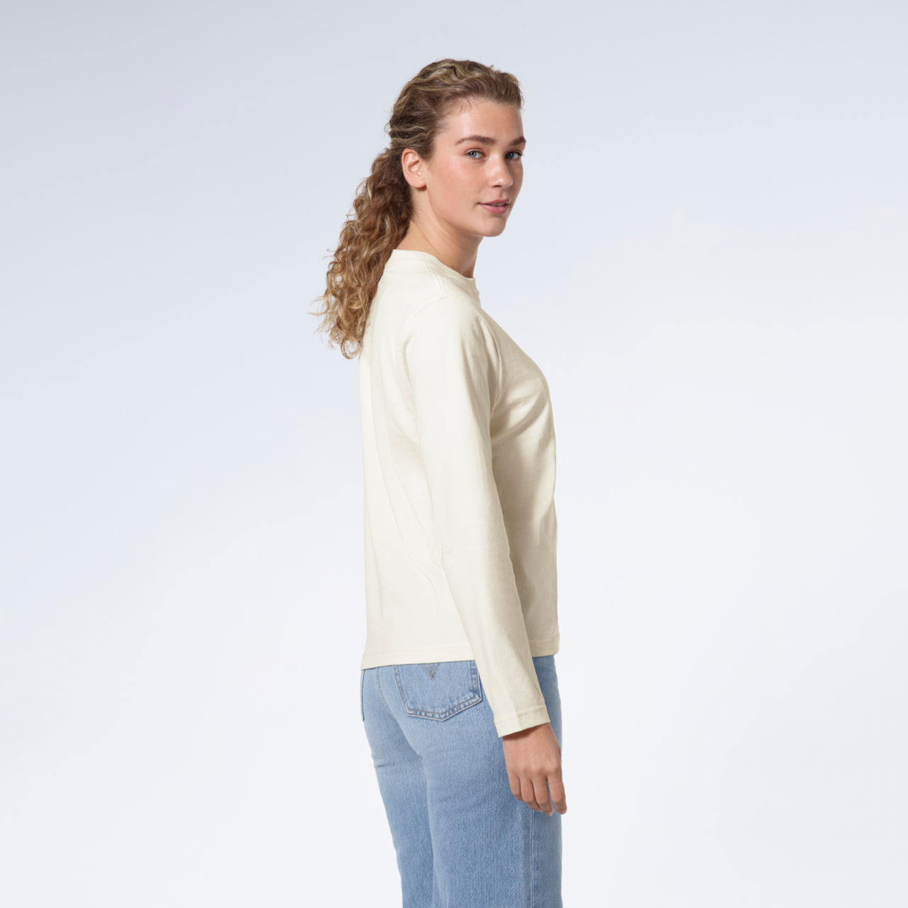 A woman facing side on looking back at the camera, modelling a plain ecru long sleeve organic cotton t shirt.