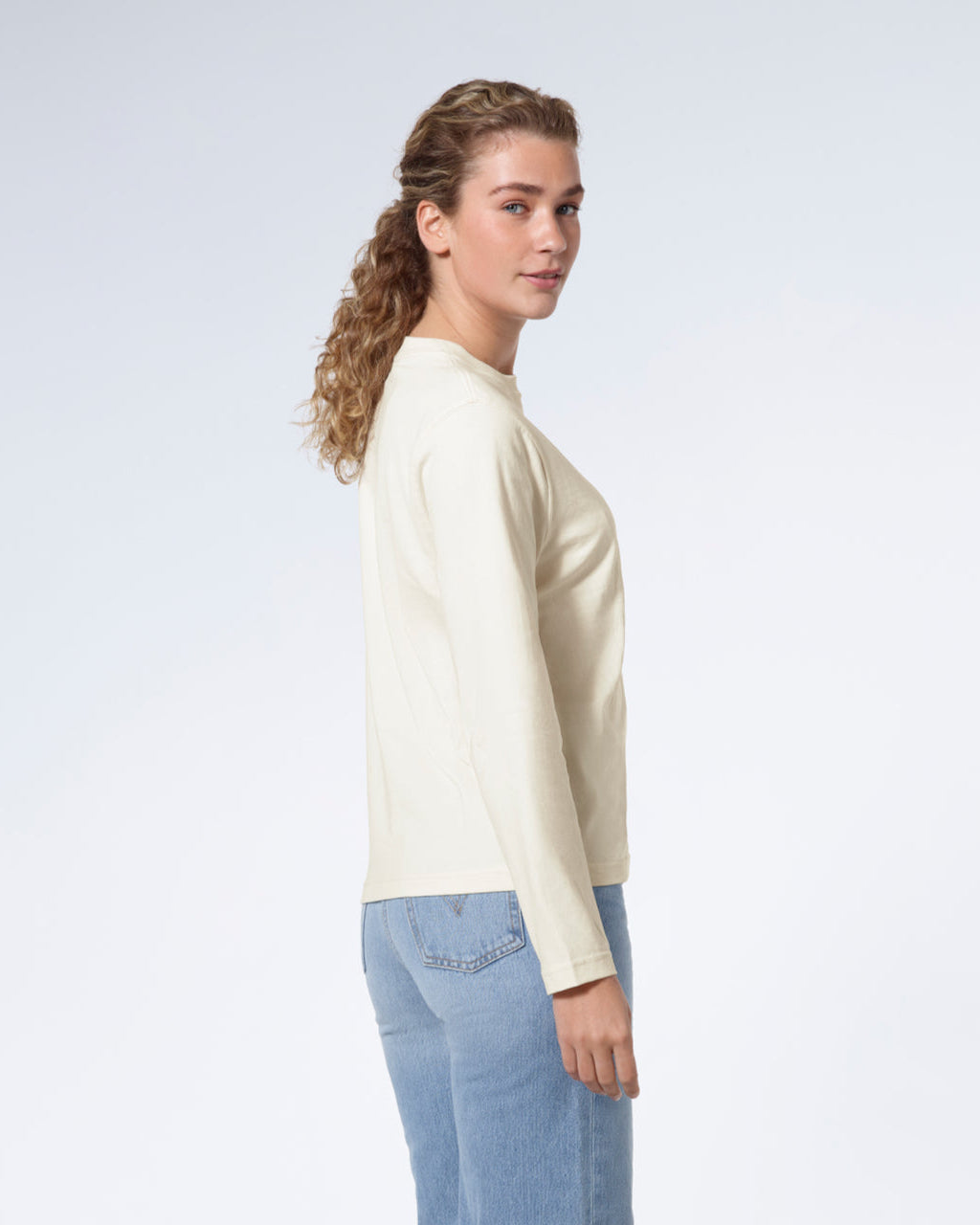 A woman facing side on looking back at the camera, modelling a plain ecru long sleeve organic cotton t shirt.