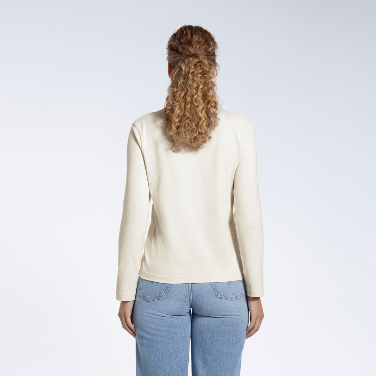 A woman facing away from the camera with her arms by her sides, modelling a plain ecru long sleeve organic cotton t shirt.