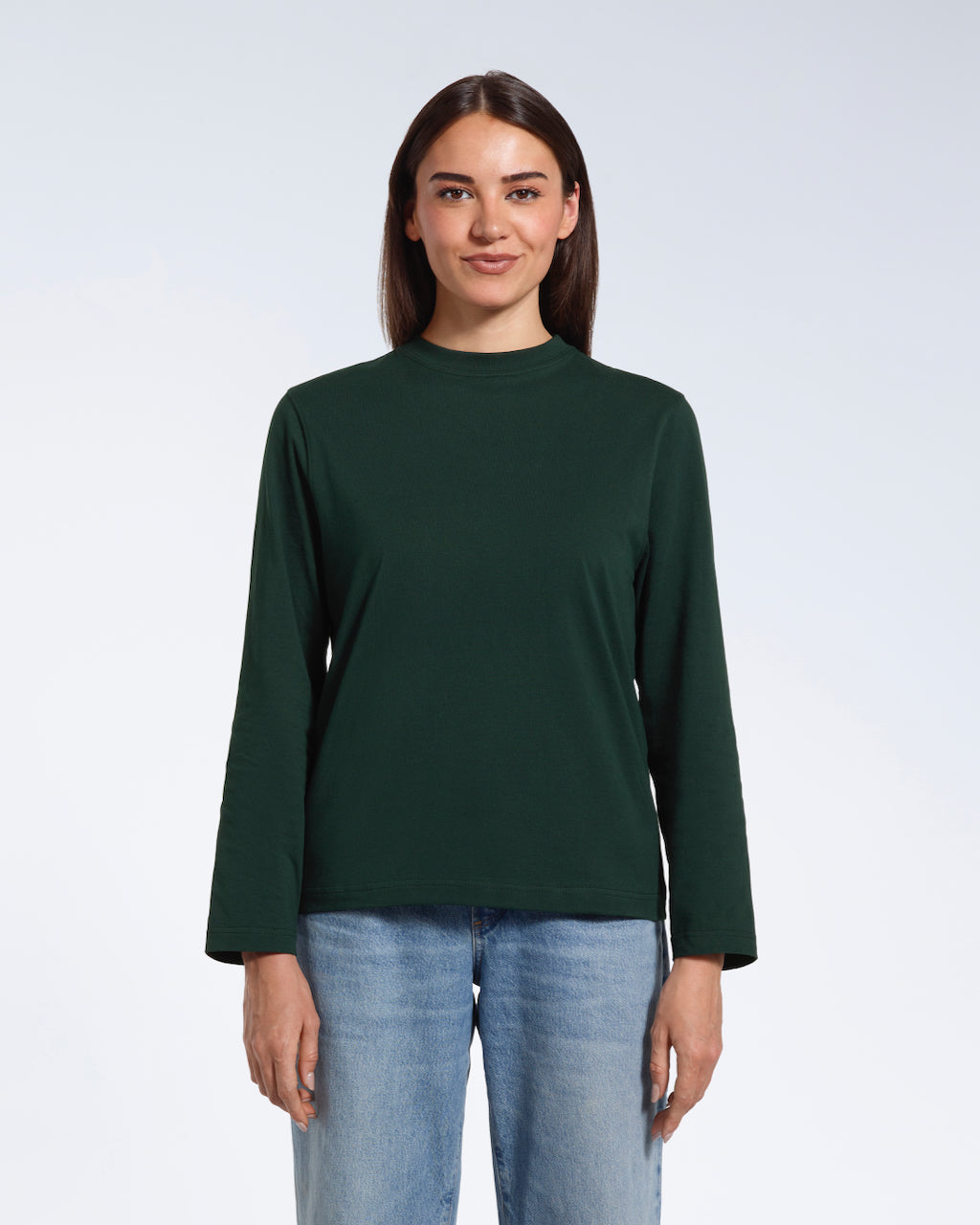 A woman standing front on with her arms by her sides, modelling a plain green long sleeve organic cotton t shirt.