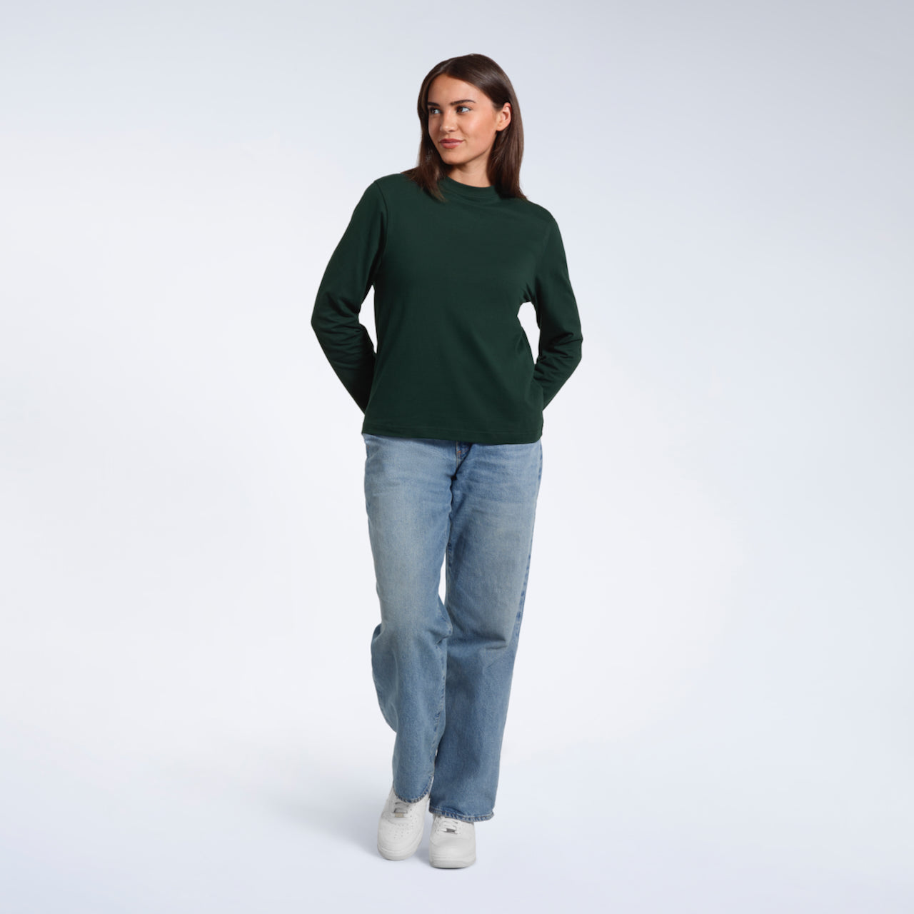 A full body photo of a woman smiling with her hands behind her back, modelling a plain green long sleeve organic cotton t shirt.