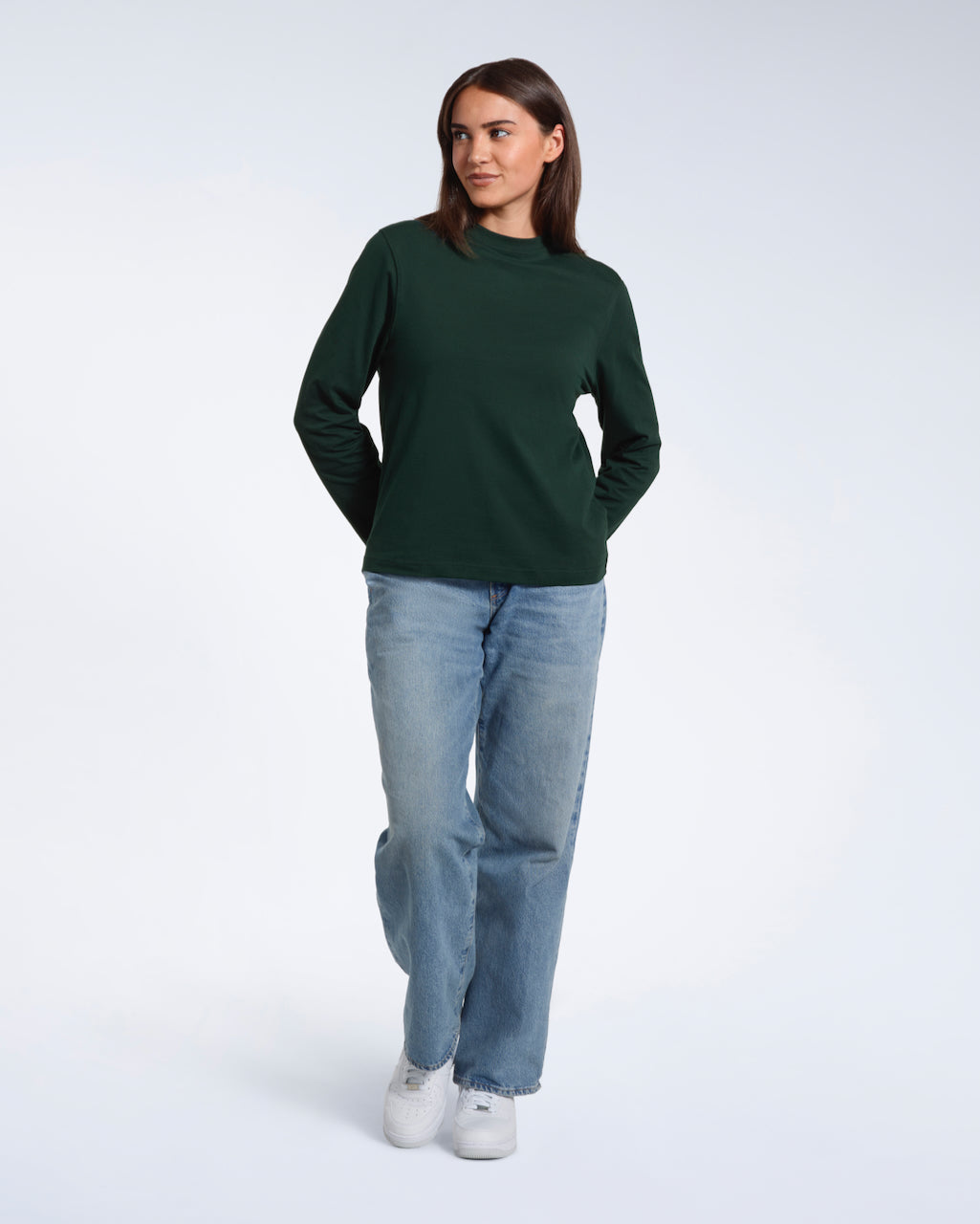 A full body photo of a woman smiling with her hands behind her back, modelling a plain green long sleeve organic cotton t shirt.