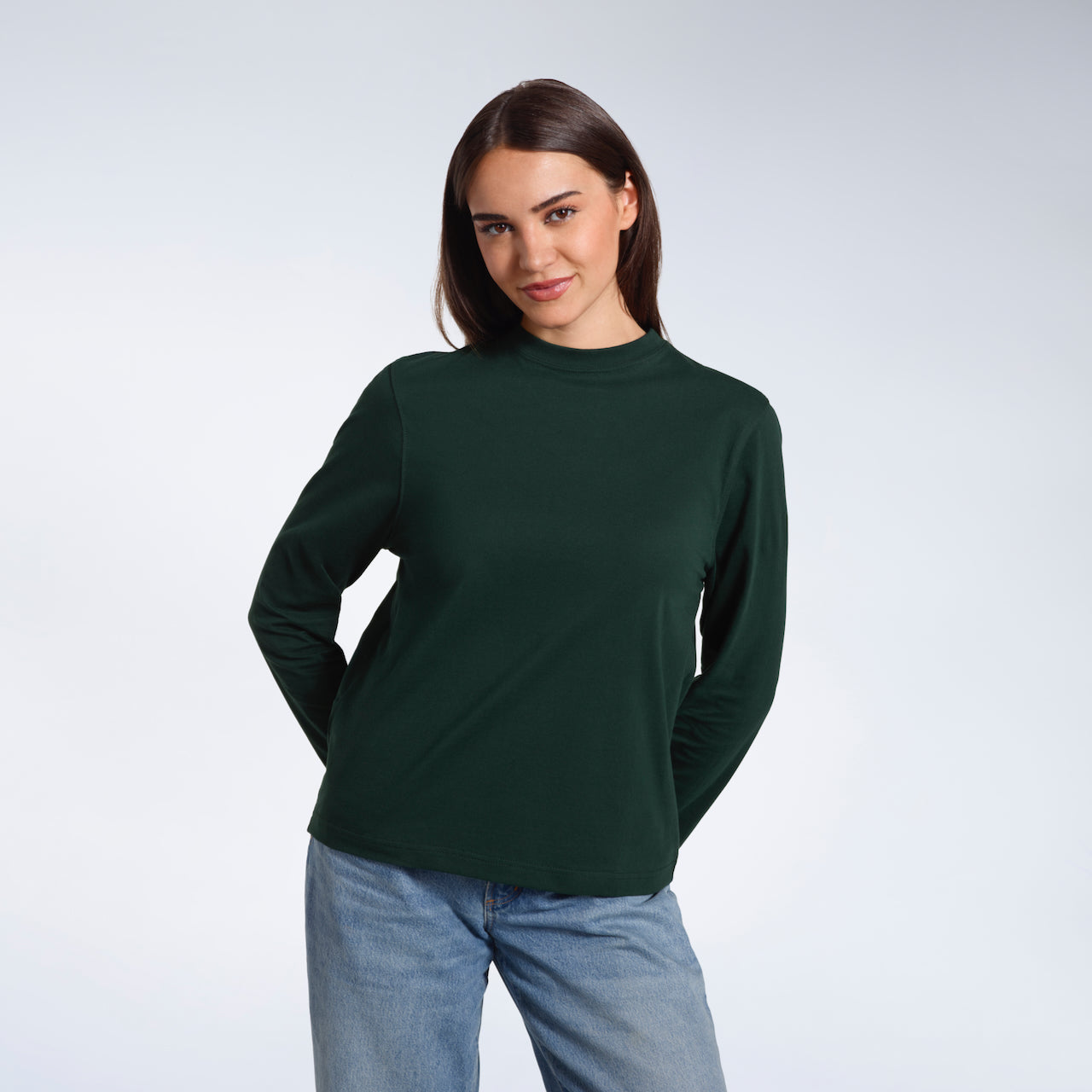 A woman looking into the camera with her hands behind her back, modelling a plain green long sleeve organic cotton t shirt.