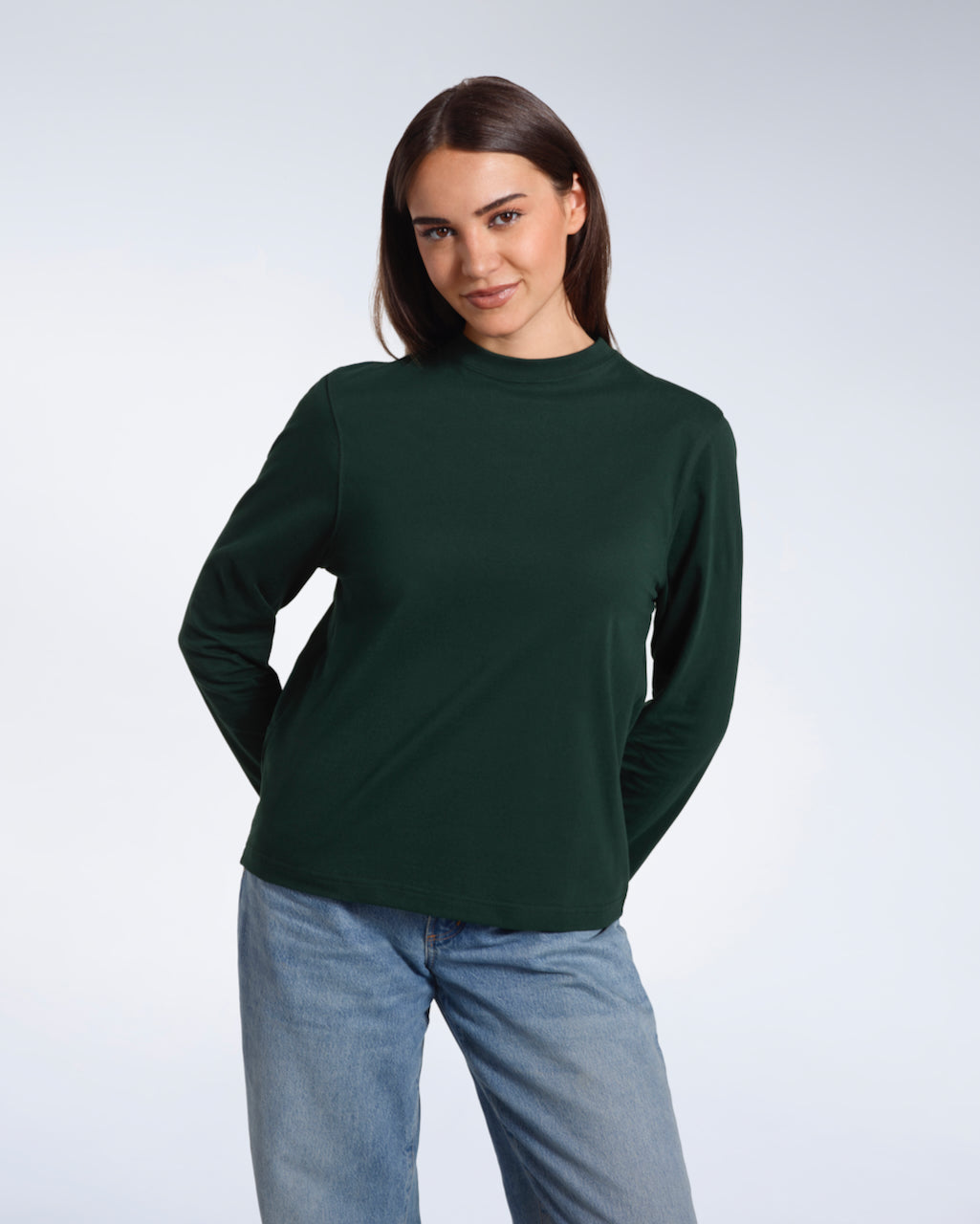 A woman looking into the camera with her hands behind her back, modelling a plain green long sleeve organic cotton t shirt.