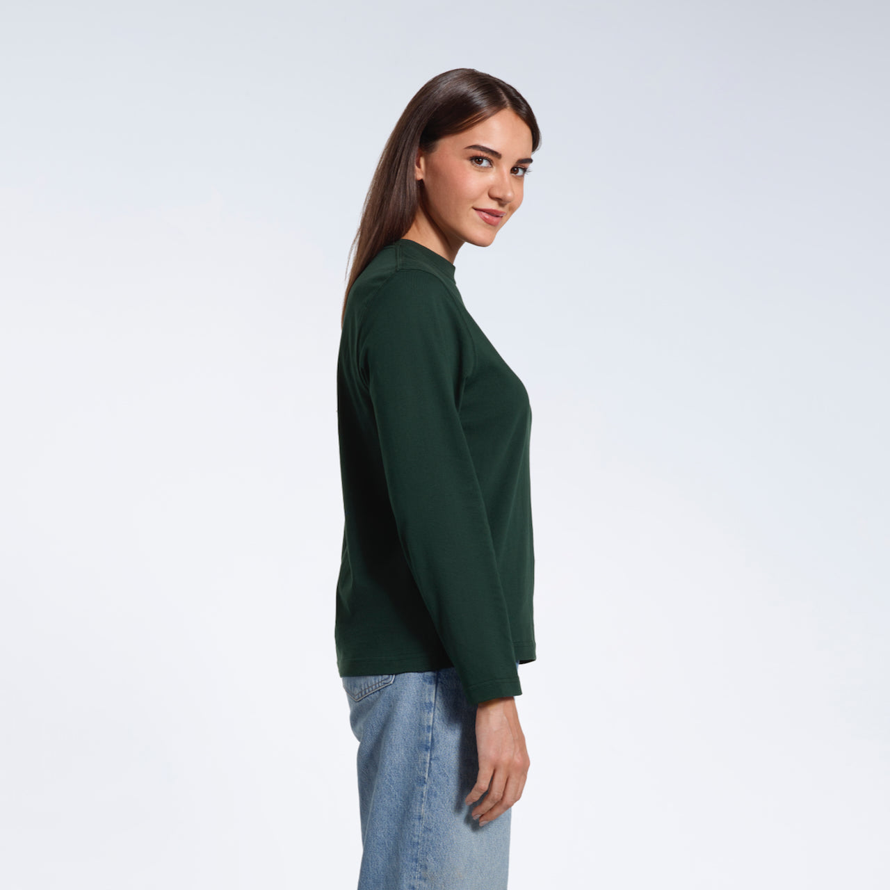 A woman facing side on looking at the camera, modelling a plain green long sleeve organic cotton t shirt.