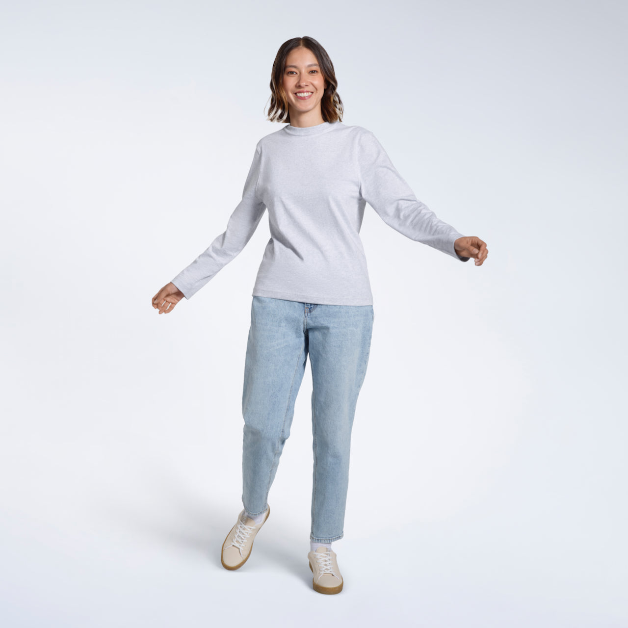 A full body photo of a woman smiling  into the camera with her arms out, modelling a plain grey long sleeve organic cotton t shirt.