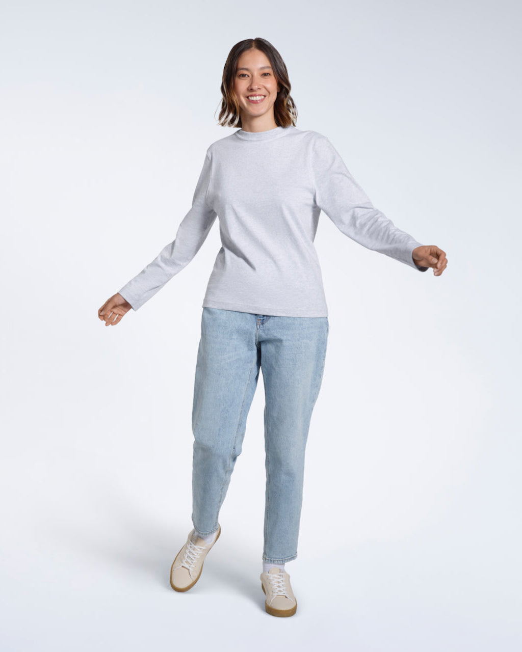 A full body photo of a woman smiling into the camera with her arms out, modelling a plain grey long sleeve organic cotton t shirt.