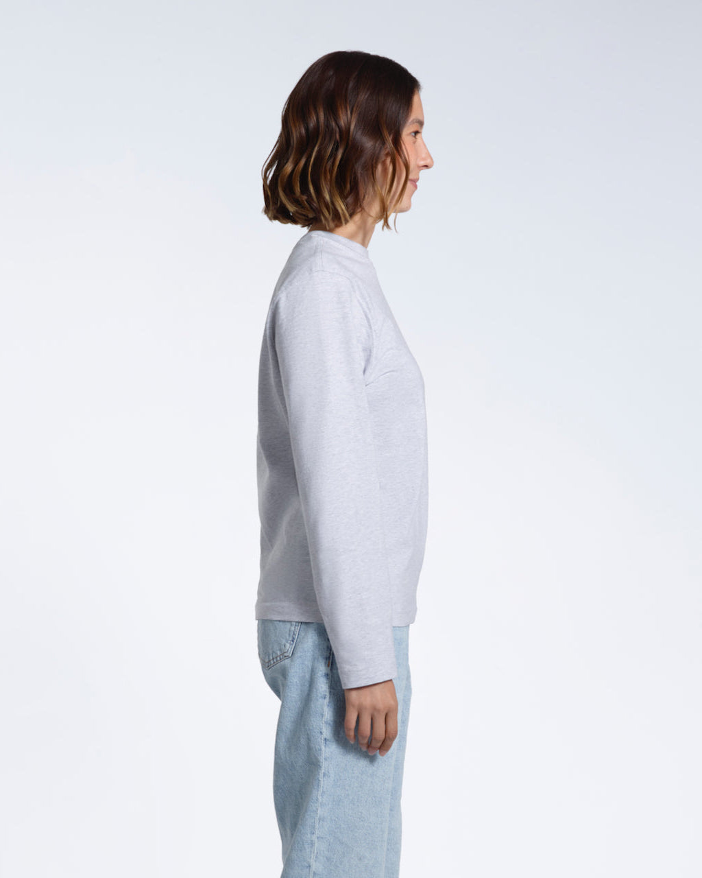 A woman facing side on with her arms by her sides, modelling a plain grey long sleeve organic cotton t shirt.