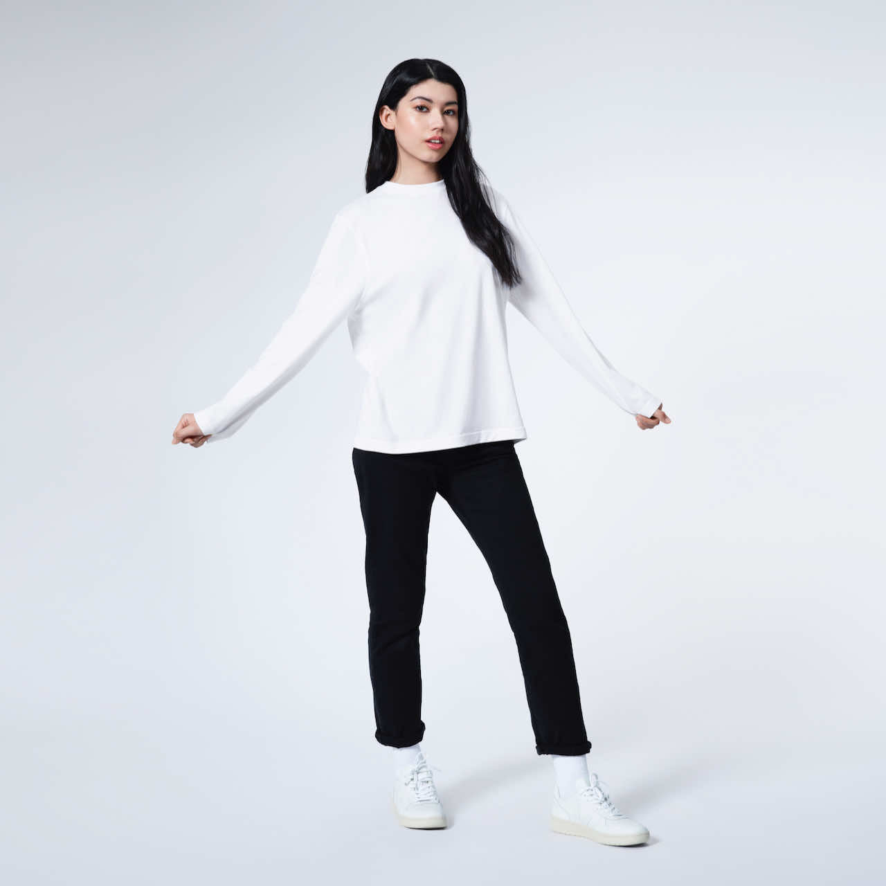 A full body photo of a woman with her arms out, modelling a plain white long sleeve organic cotton t shirt.