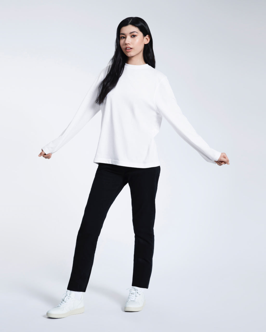 A full body photo of a woman with her arms out, modelling a plain white long sleeve organic cotton t shirt.
