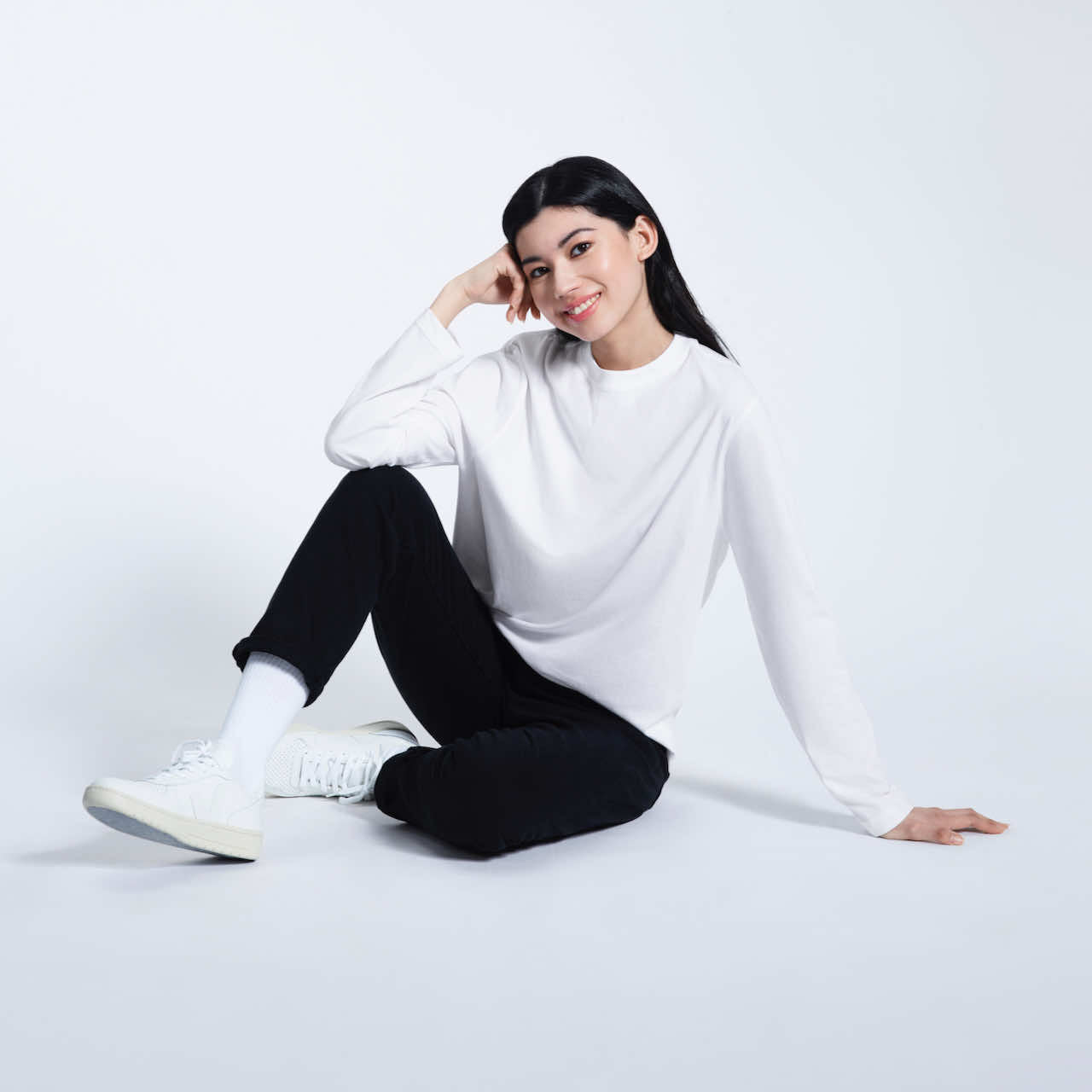 A woman sitting on the floor smiling, modelling a plain white long sleeve organic cotton t shirt.