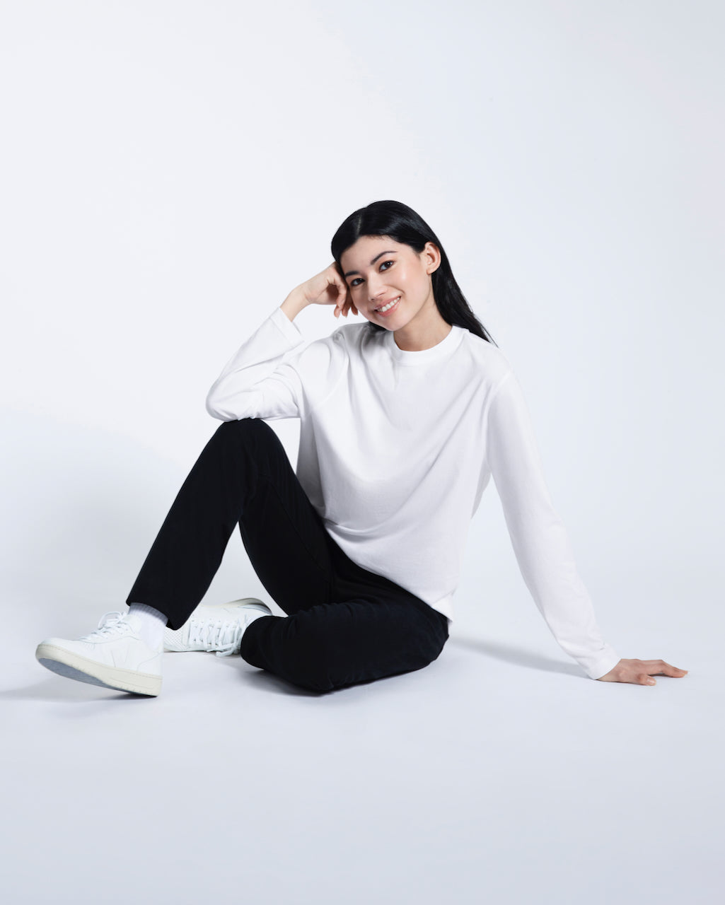 A woman sitting on the floor smiling, modelling a plain white long sleeve organic cotton t shirt.