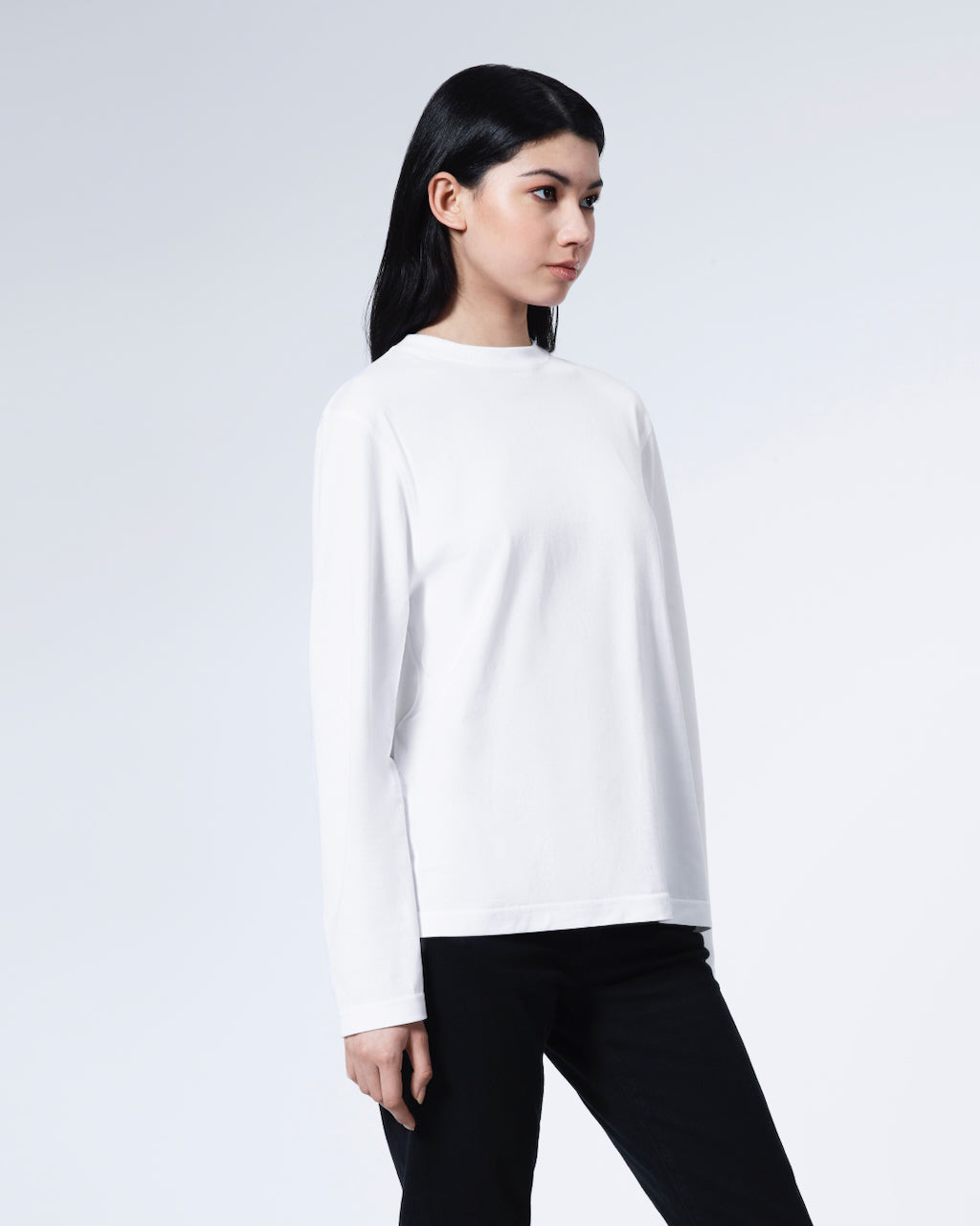 A woman looking away from the camera with her arms by her sides, modelling a plain white long sleeve organic cotton t shirt.