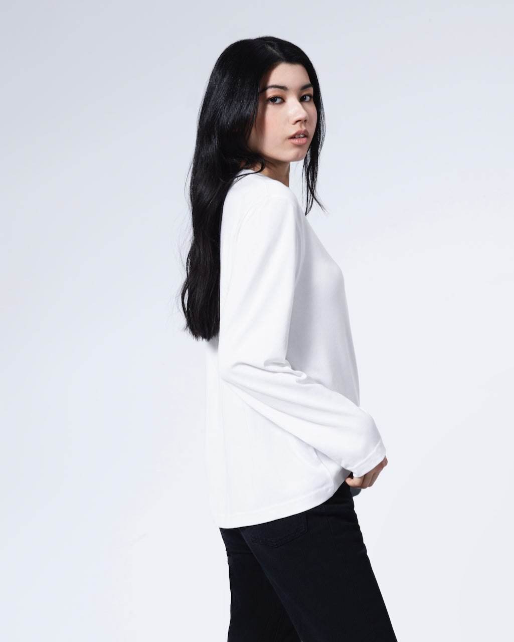 A woman facing side looking back at the camera, modelling a plain white long sleeve organic cotton t shirt.