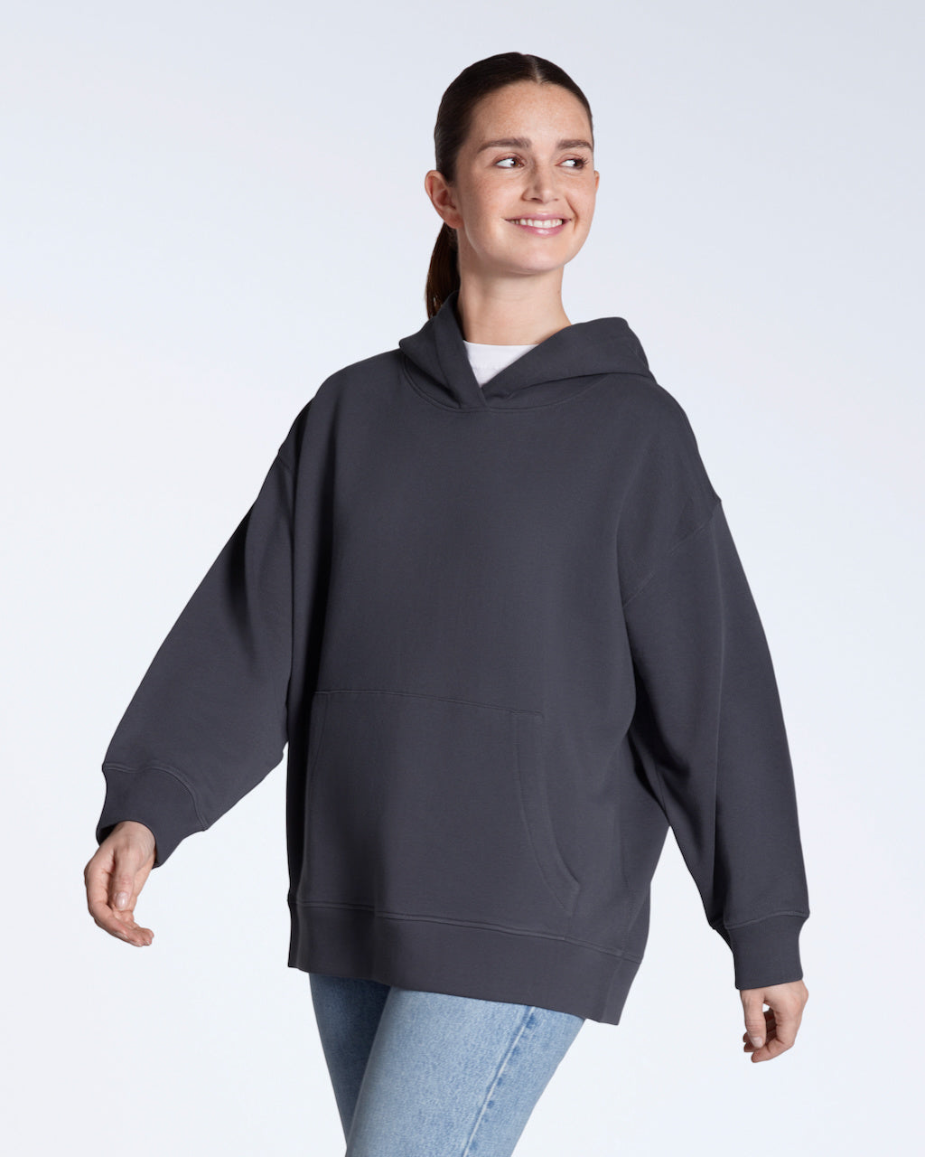 A woman walking and amiling, modelling a plain dark grey, oversized, organic cotton hoodie.