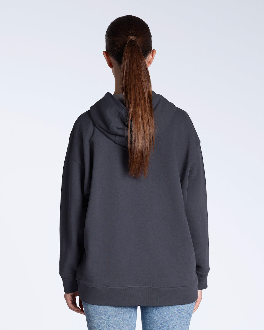 A woman facing away from the camera with her hands by her sides, modelling a plain dark grey, oversized, organic cotton hoodie.