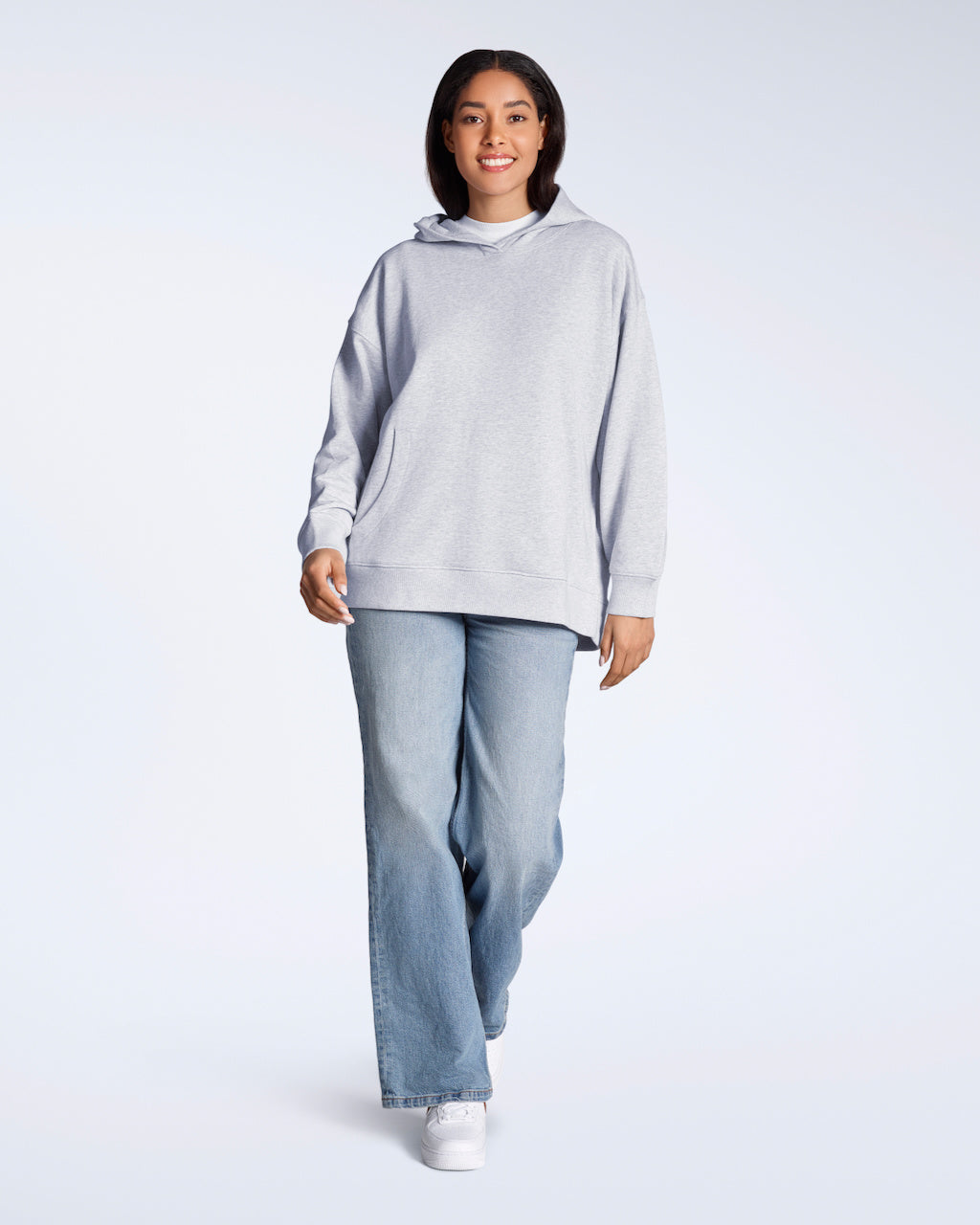 A full body photo of a woman smiling into the camera, modelling a grey, oversized, organic cotton hoodie with a pair of blue jeans.
