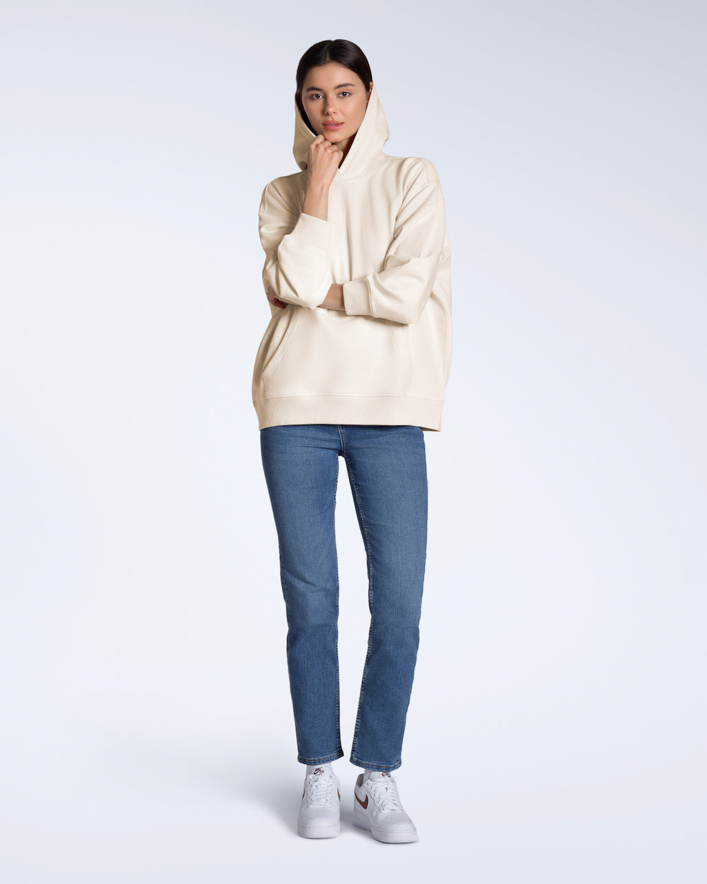 A full body photo of a woman looking into the camera touching her hood, modeling a plain, natural coloured, oversized, organic cotton hoodie.