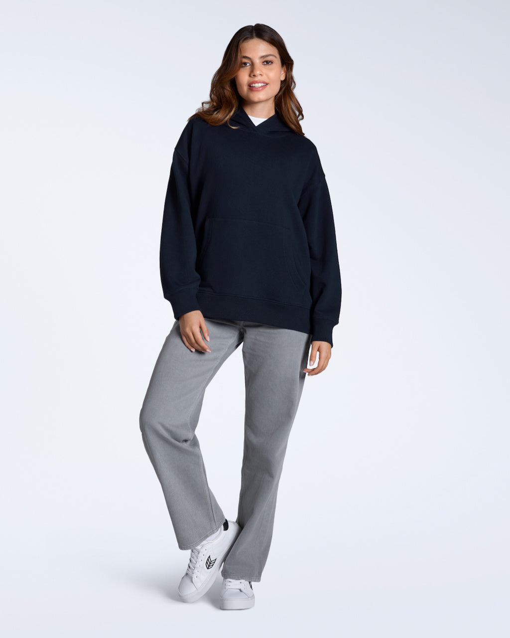 A full body photo of a woman smiling into the camera, modelling a plain navy, oversized, organic cotton hoodie with a pair of grey trousers.