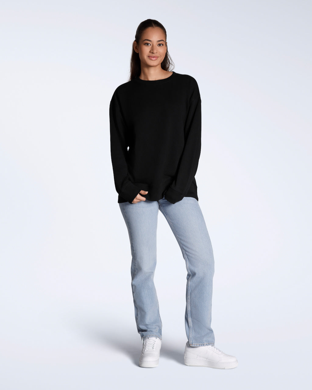 A full body photo of a woman smiling into the camera, modelling a plain black, oversized, organic cotton sweatshirt.