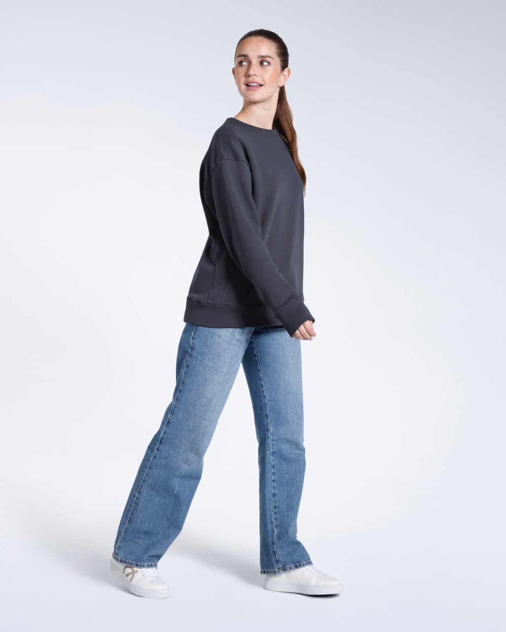 A full body photo of a woman looking over her shoulder, modelling a plain dark grey oversized organic cotton sweatshirt.