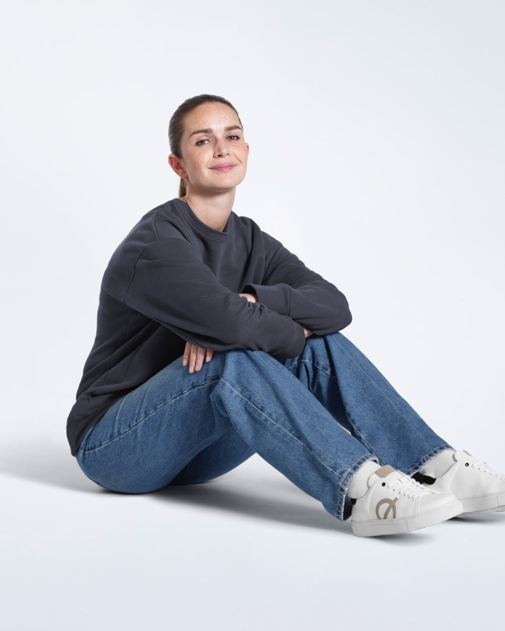 A woman sitting on the floor smiling, modelling a plain dark grey oversized organic cotton sweatshirt.
