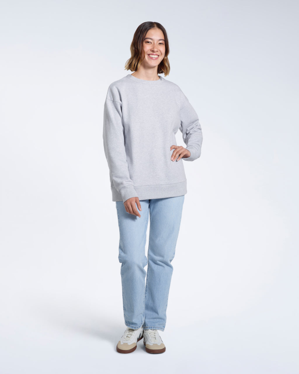 A full body photo of a woman smiling with her hand on her hip, modelling a plain grey oversized organic cotton sweatshirt.