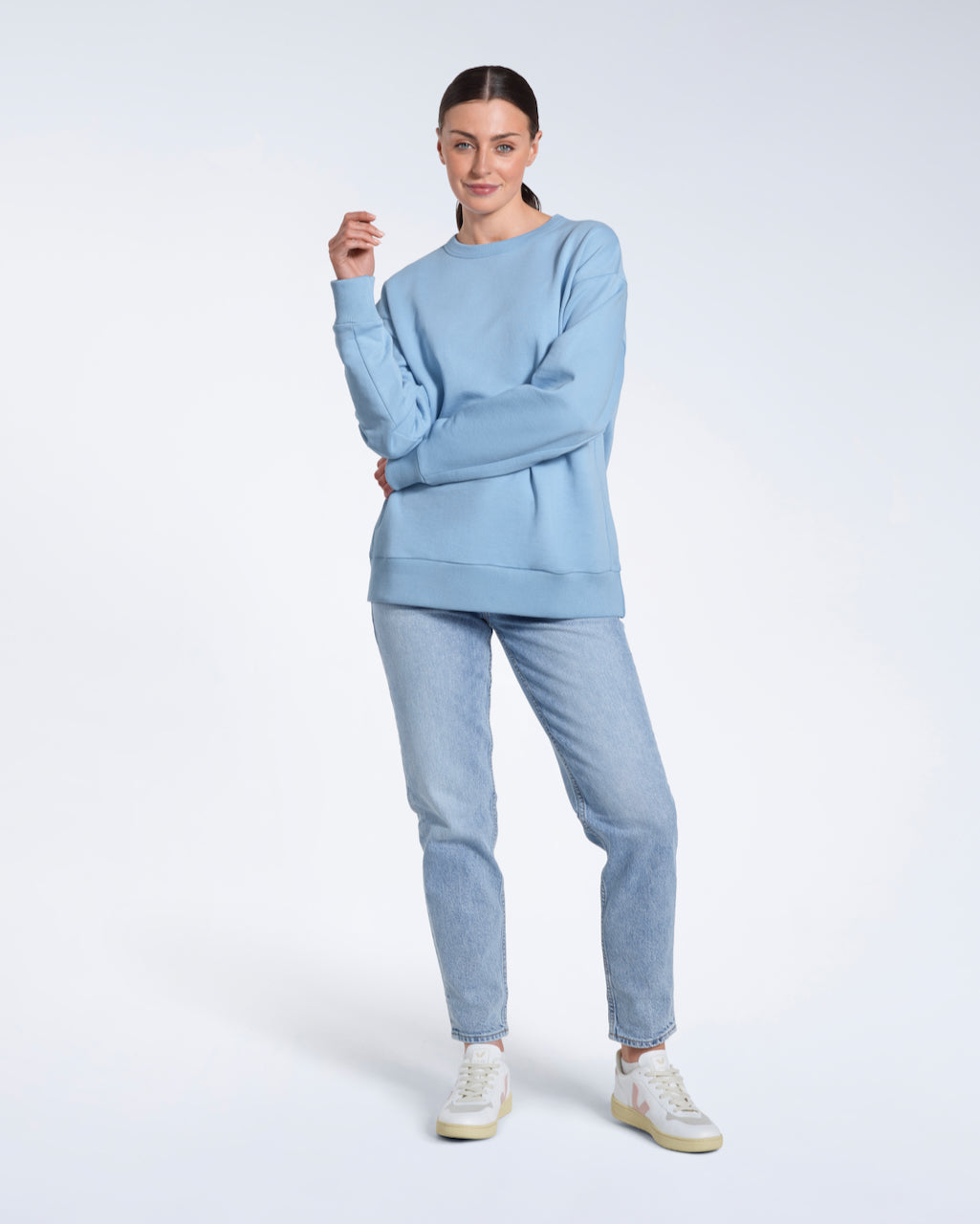 A full body photo of a woman smiling into the camera with her arm across her body, modelling a plain blue oversized organic cotton sweatshirt.