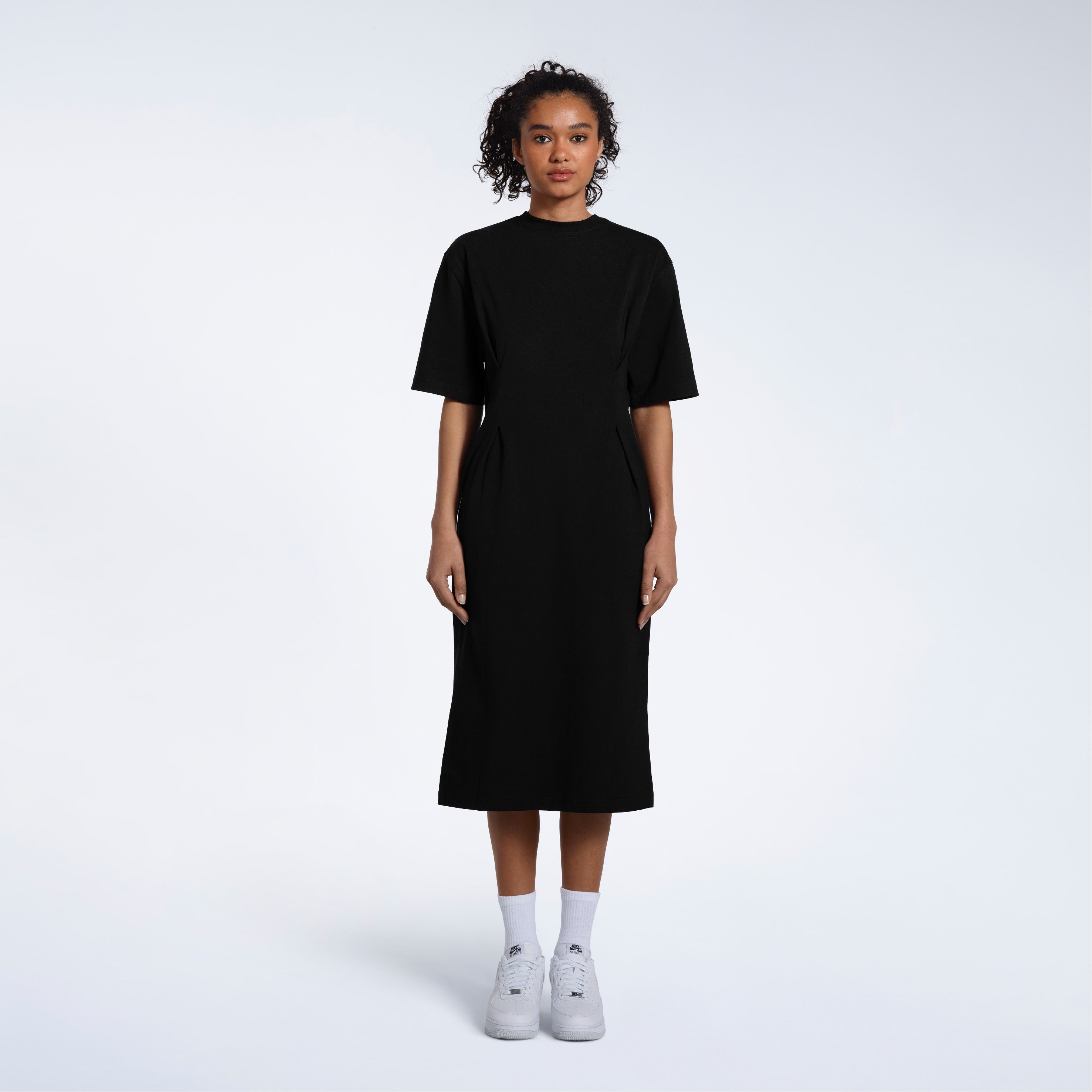 A women standing face on with her arms by her sides, modelling a plain black, organic cotton midi t shirt dress.