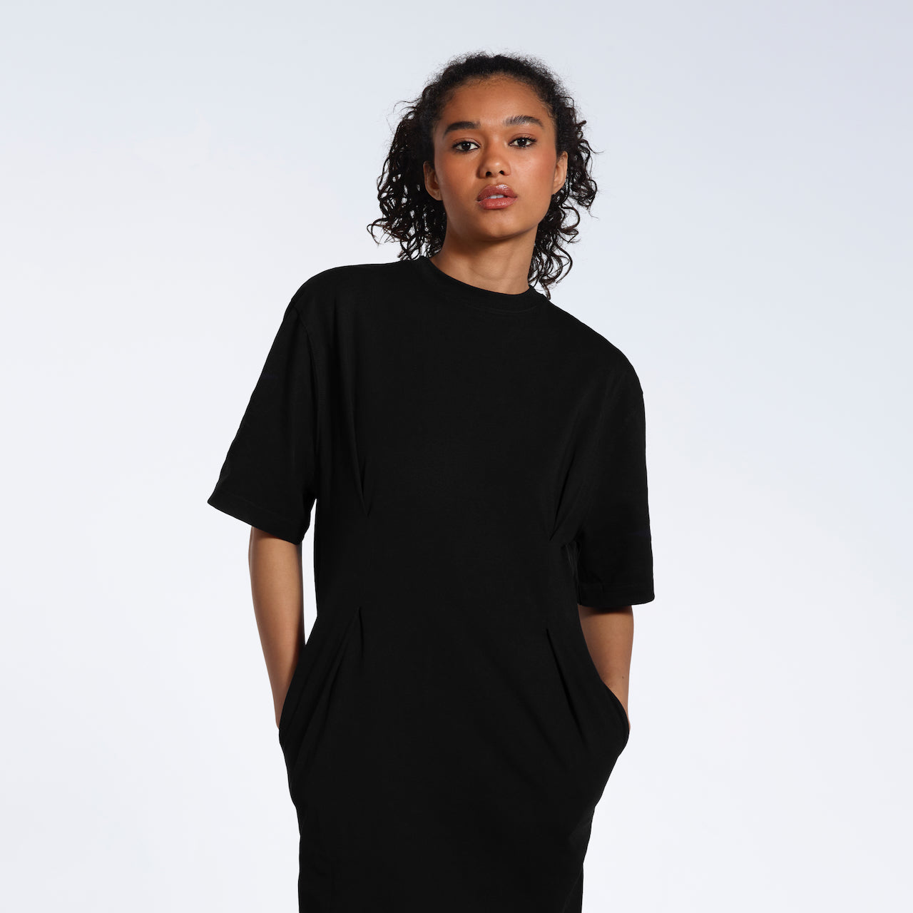 A women looking into the camera with her hands in her pockets, modelling a plain black, organic cotton midi t shirt dress.