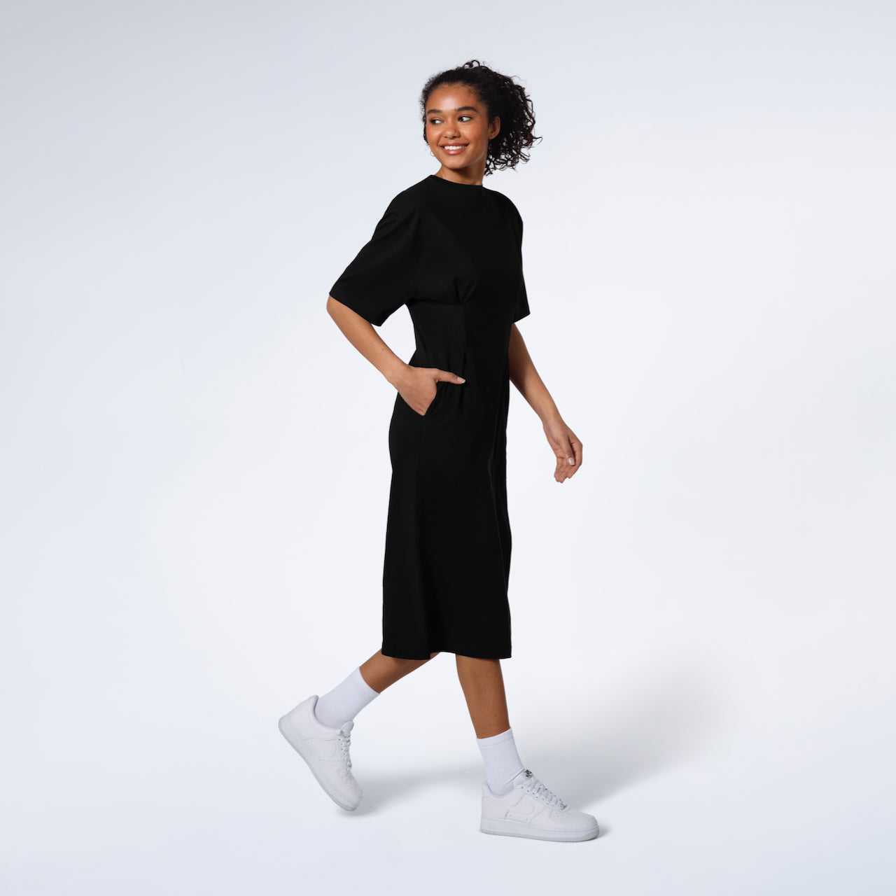 A women smiling and walking with her hand in her pocket, modelling a plain black, organic cotton midi t shirt dress.