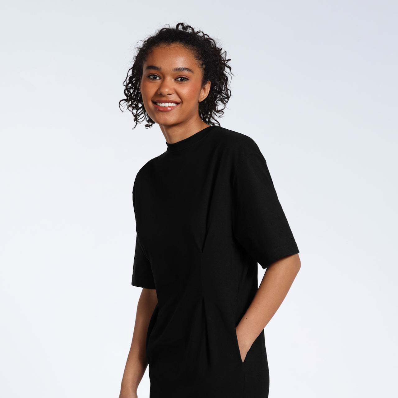 A women looking into the camera smiling with her hand in her pocket, modelling a plain black, organic cotton midi t shirt dress.