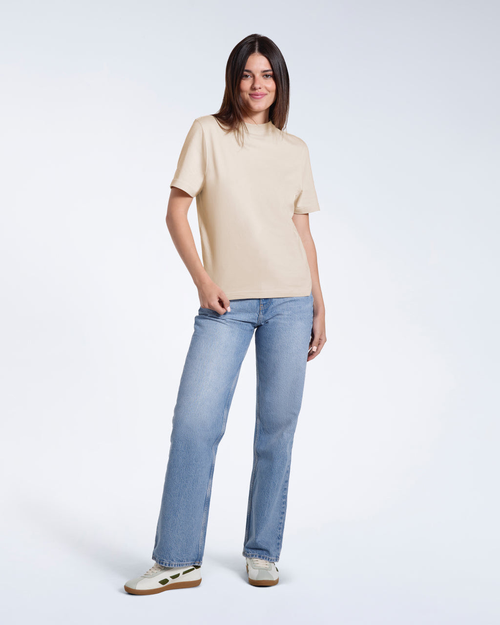 A full body photo of a woman looking into the camera smiling, modelling a plain beige short sleeve organic cotton t shirt.