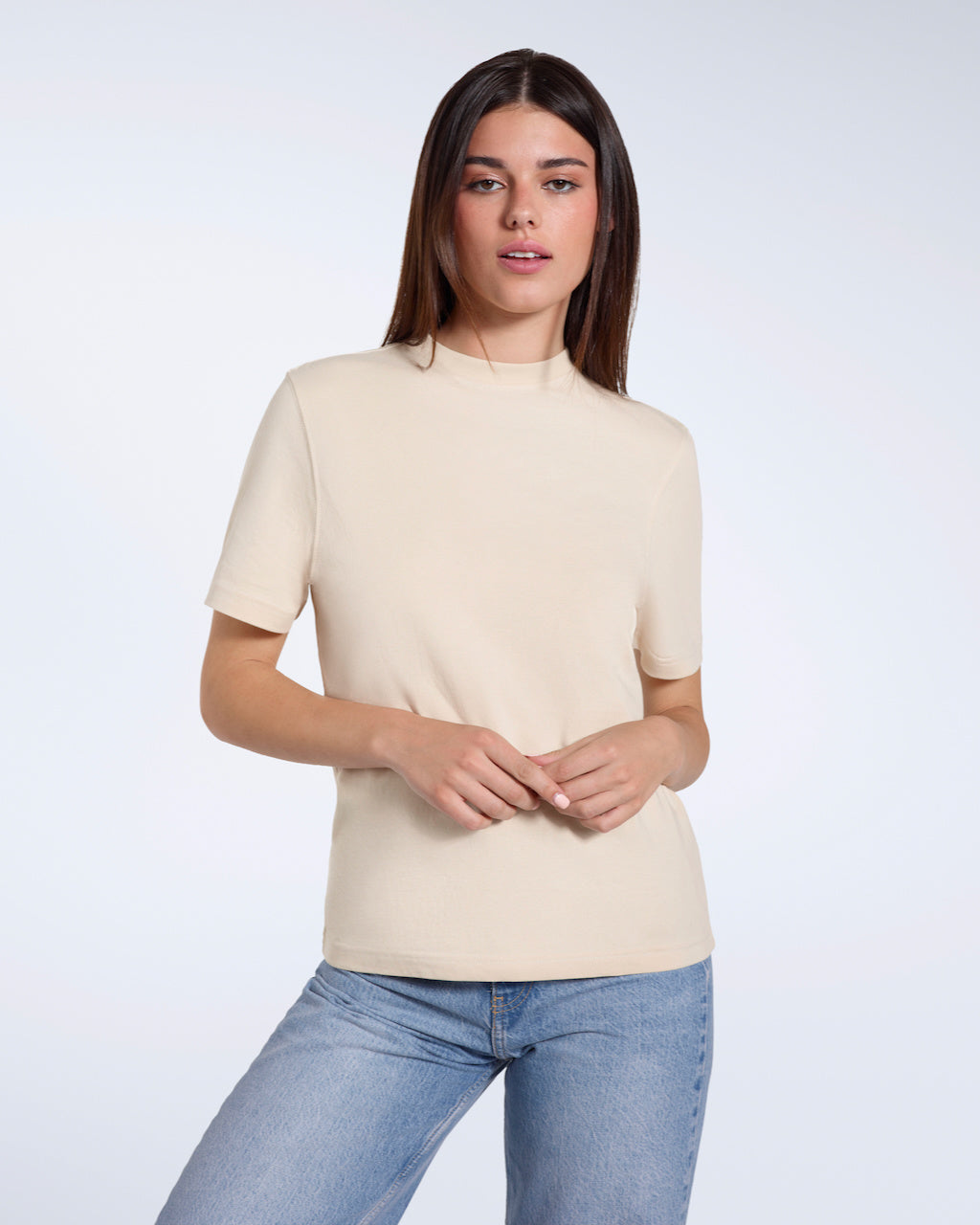 A woman with her hands above her head looking into the camera, modelling a plain beige short sleeve organic cotton t shirt.