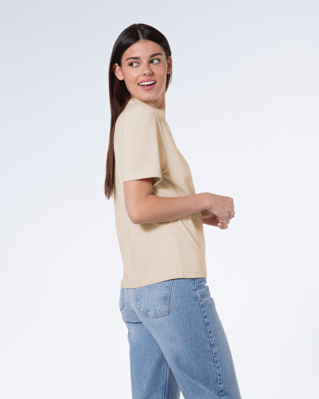 A woman facing side on and looking behind her smiling, modelling a plain beige short sleeve organic cotton t shirt.