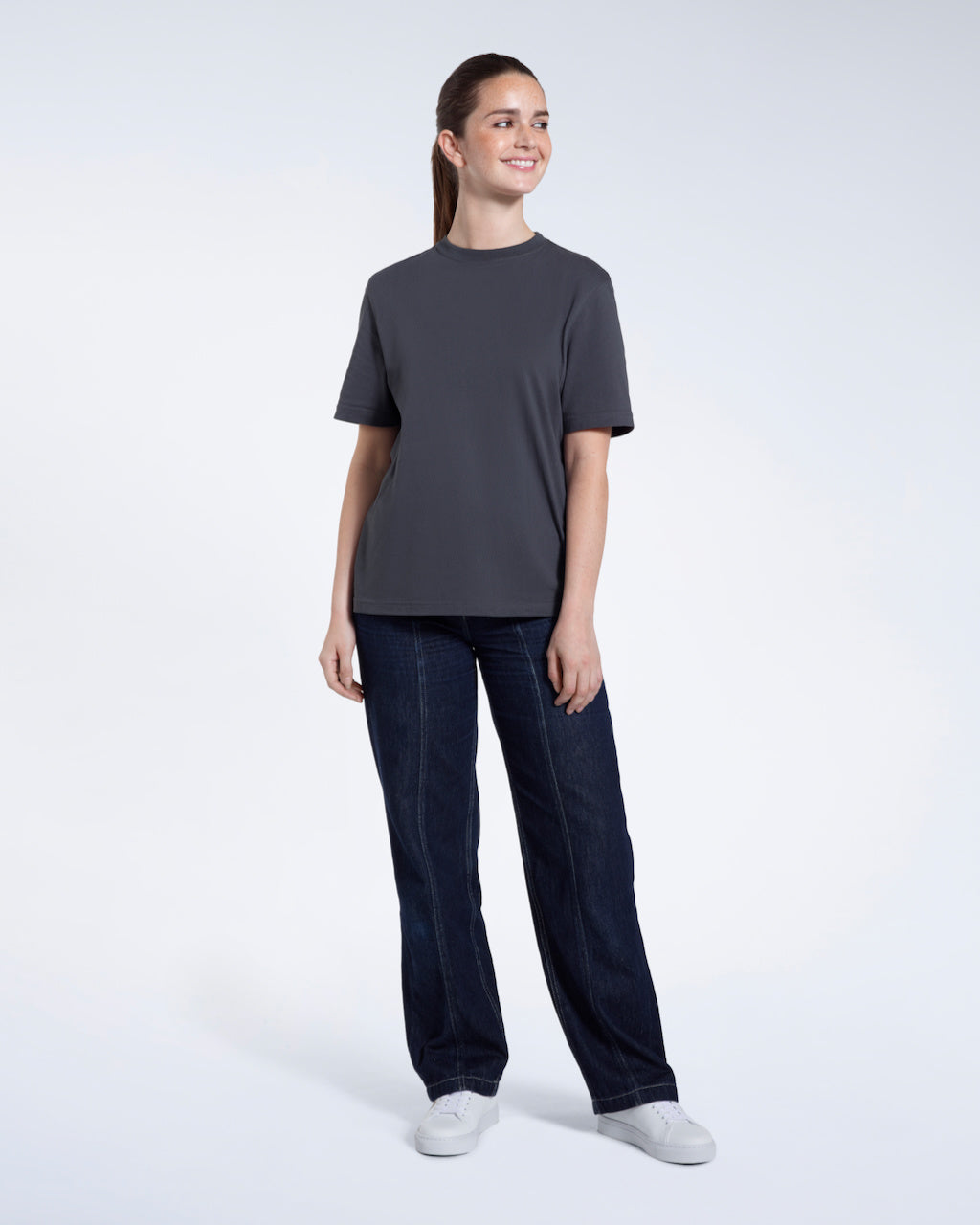 A full body photo of a woman smiling with her arms by her sides, modelling a plain dark grey short sleeve organic cotton t shirt.
