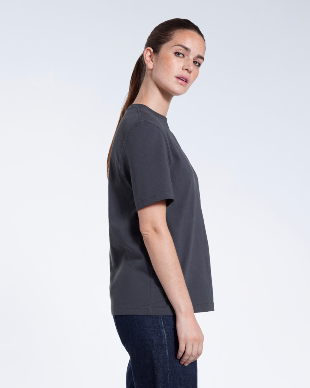 A woman facing side on and looking at the camera, modelling a plain dark grey short sleeve organic cotton t shirt.