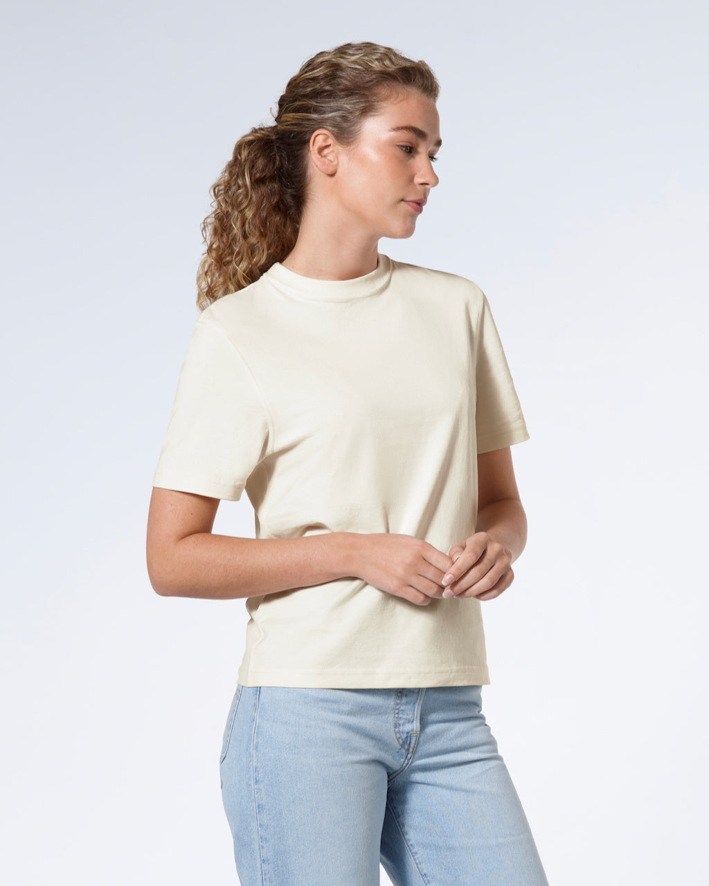 A woman with her hands together looking away from the camera, modelling a plain ecru short sleeve organic cotton t shirt.